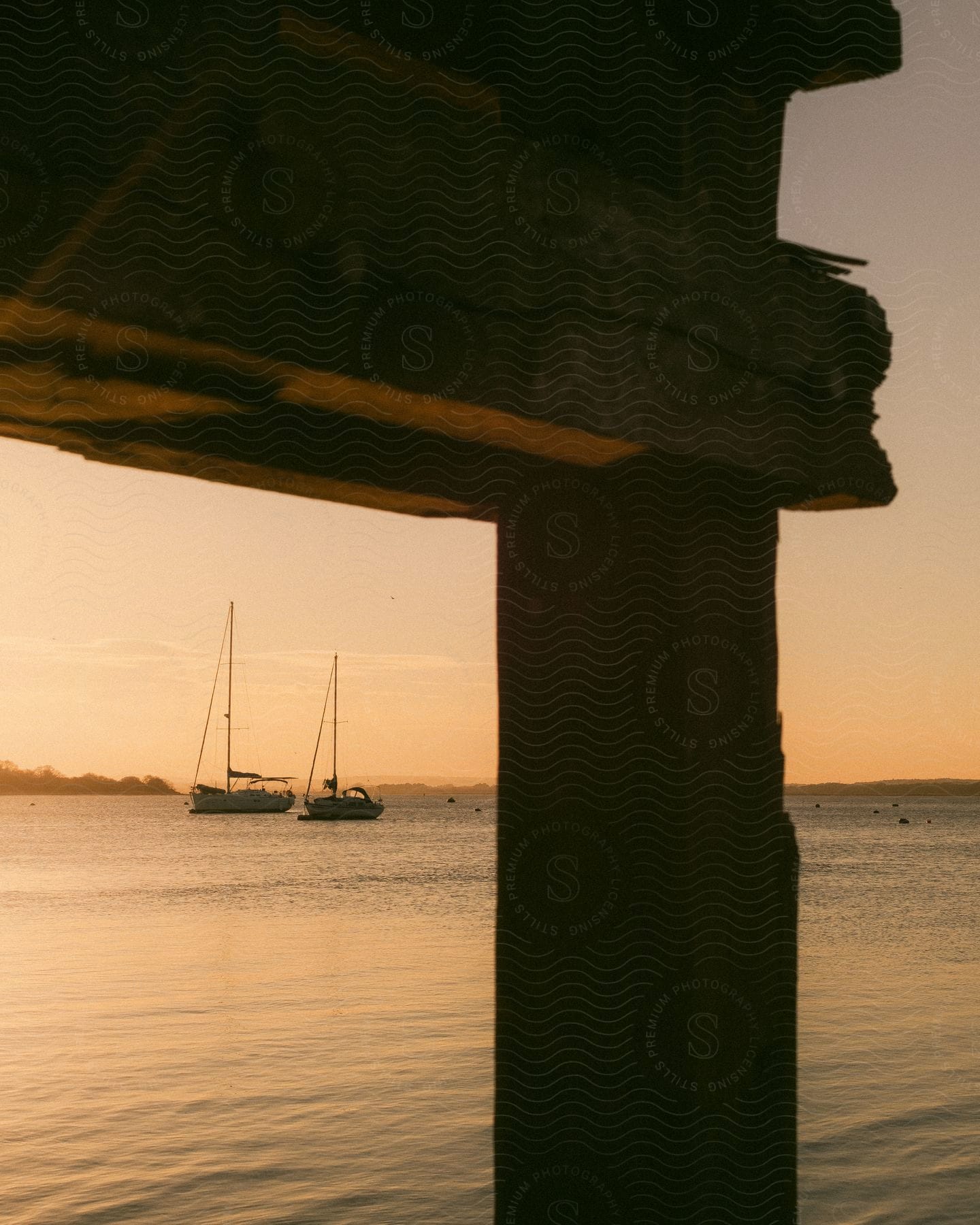 A serene sunset scene in a harbor, with two sailboats in the distance, framed by part of an aged wooden structure.
