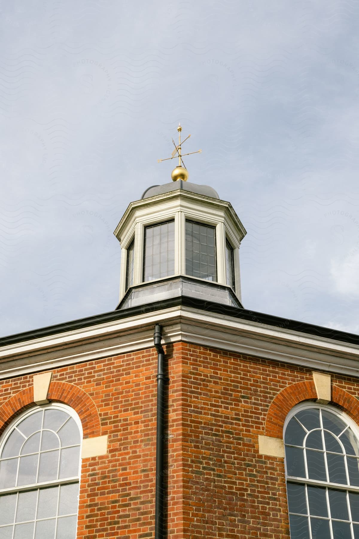 Top of a brick building with windows and a small domed tower and a golden weather vane against a blue sky with clouds.