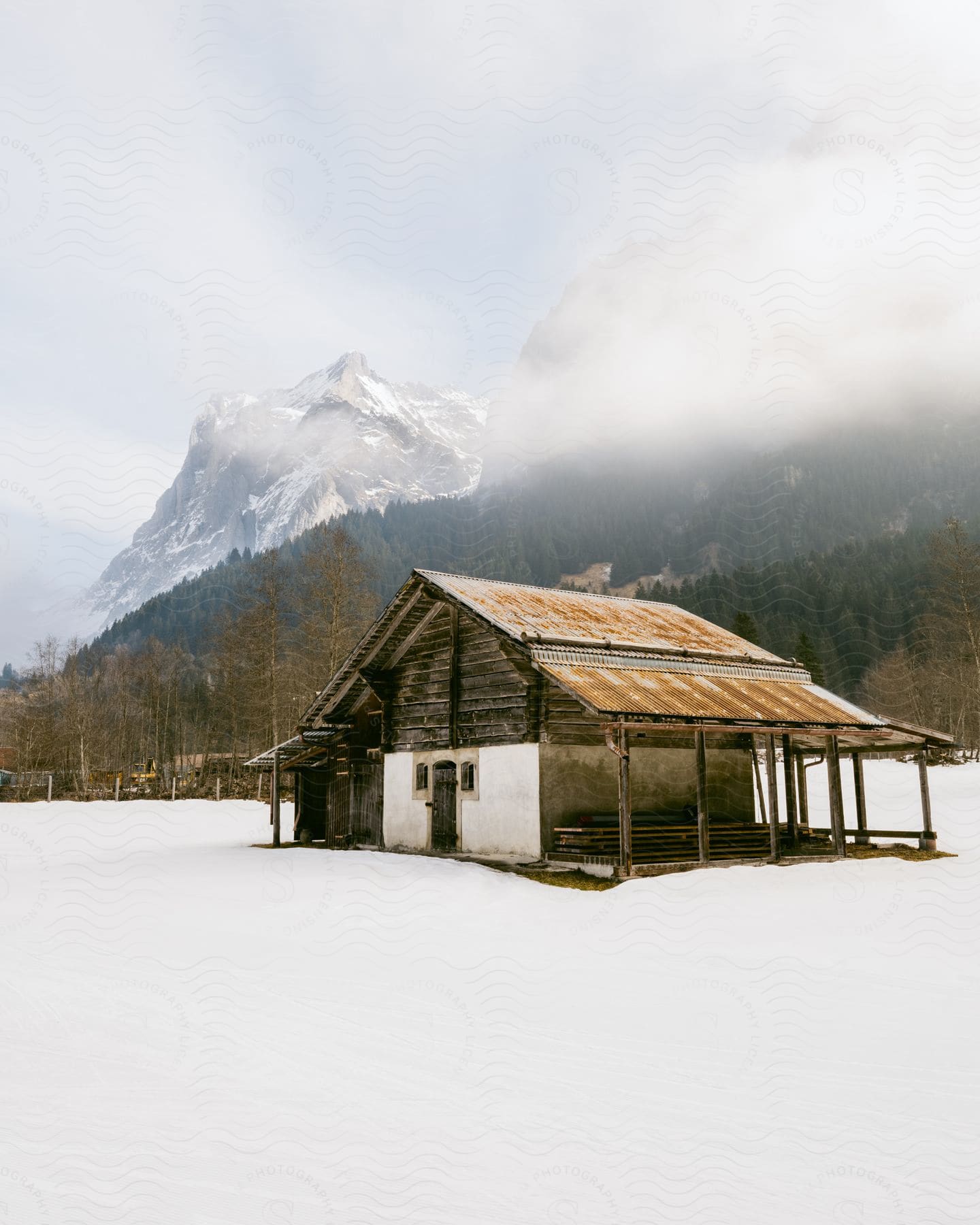 A rustic cabin rests peacefully in the middle of a snowy landscape, with a majestic mountain shrouded in mist in the background.