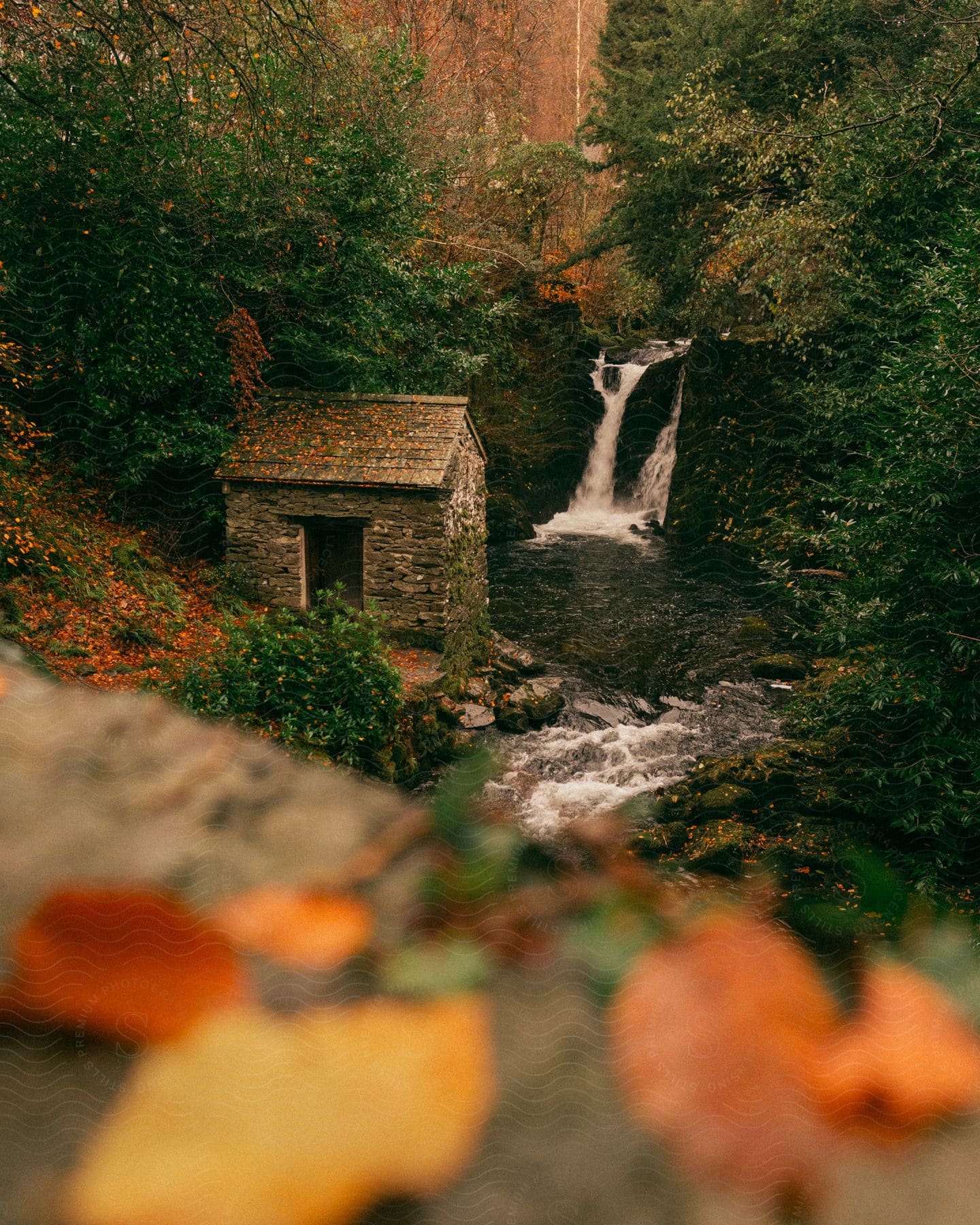 A stone hut sits on a rocky bank next to a small waterfall on an autumn day.