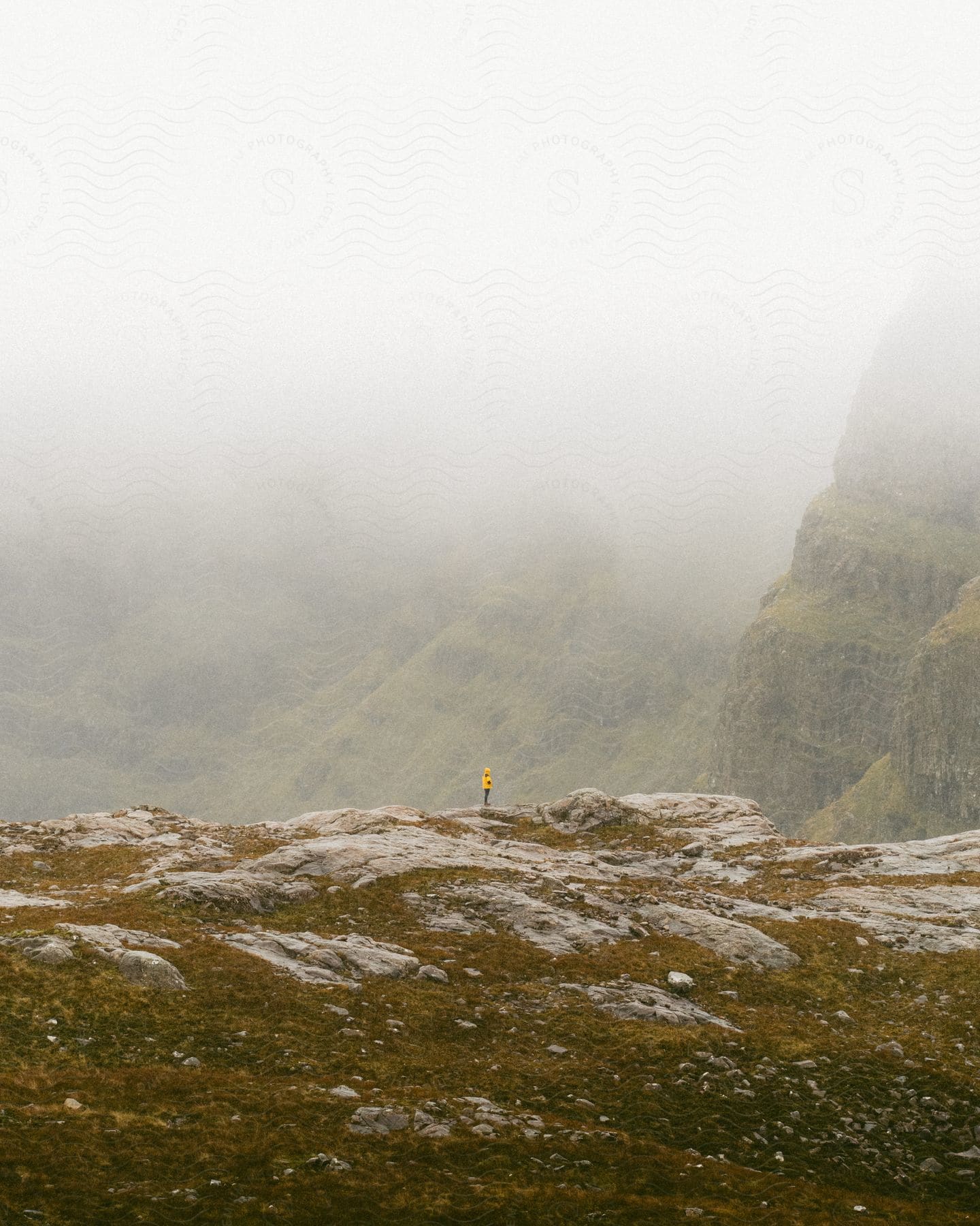 Person in yellow jacket standing on rocky mountain plateau, misty hills in background.