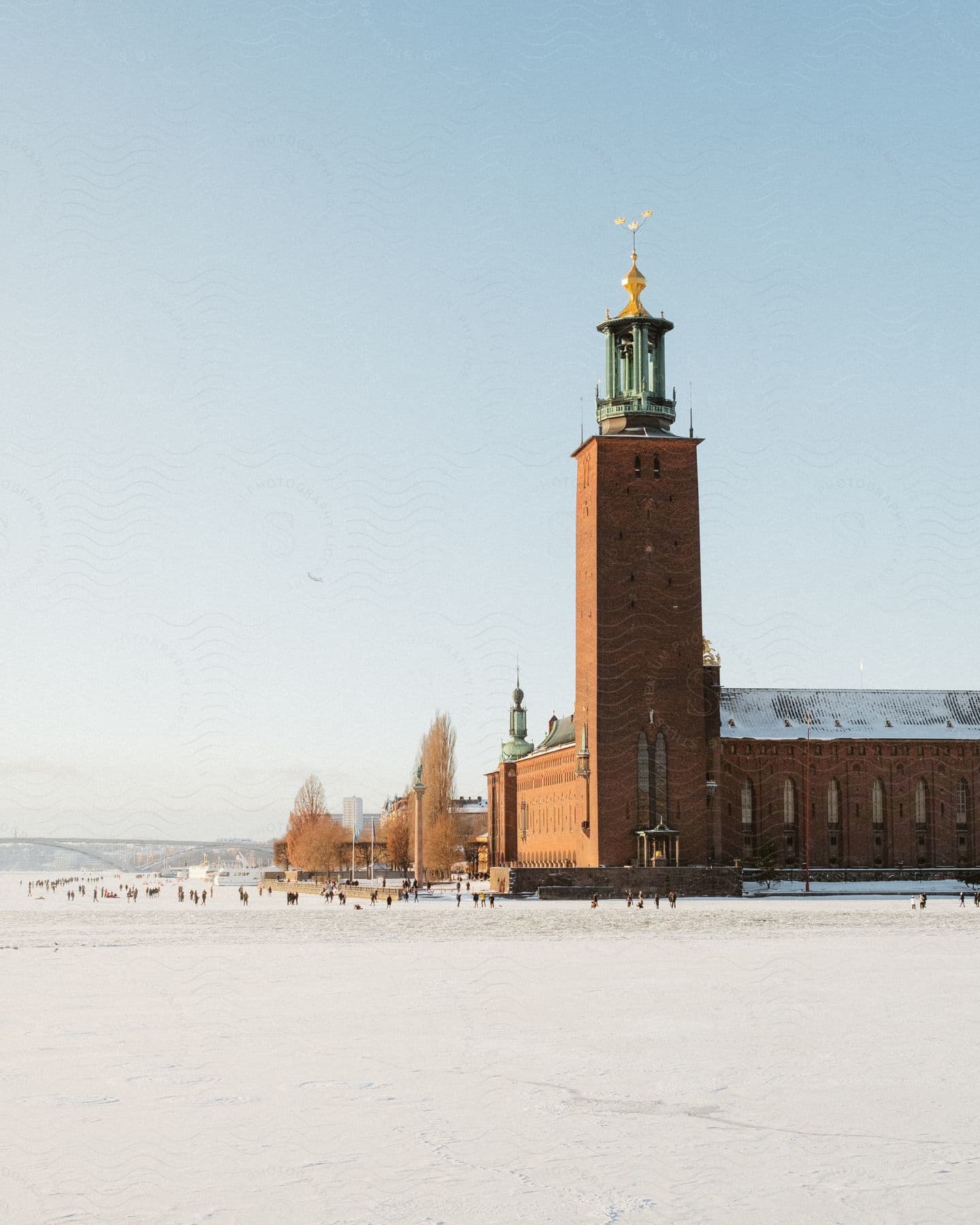 A brick building has a tower with a very ornate spire atop it and is surrounded by people standing on snow.