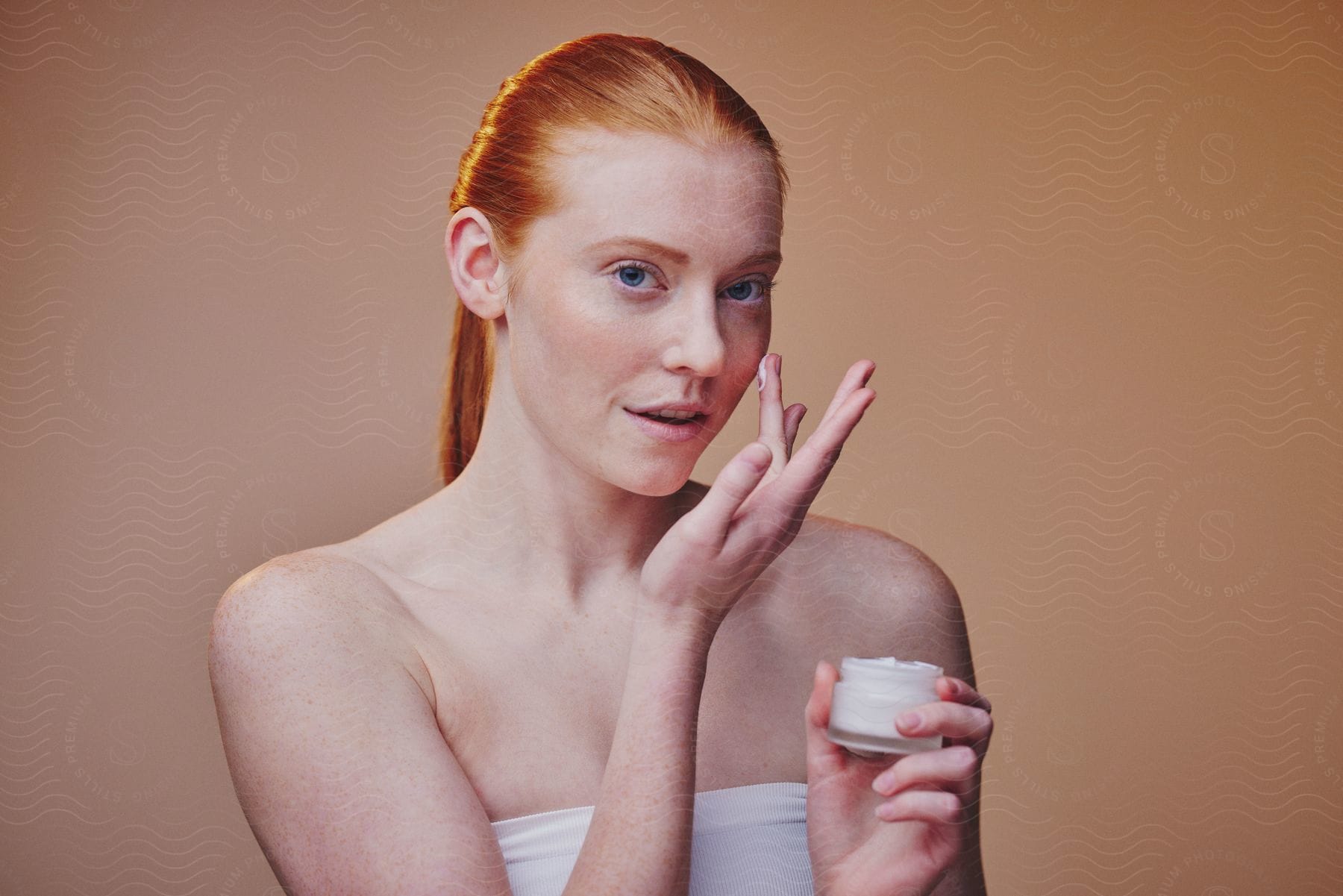 A woman with fiery red hair, wearing a white top, applies cream to her face from a jar she holds in her hand.