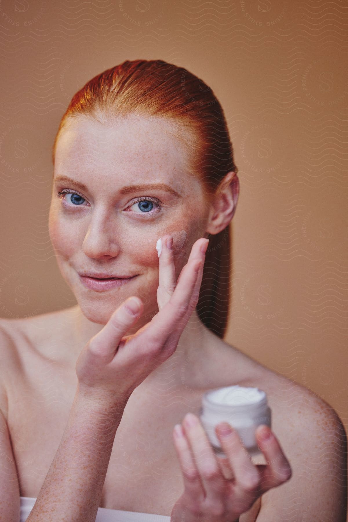 Red-haired woman applying cream to her face with her fingers and holding the bottle with her other hand.