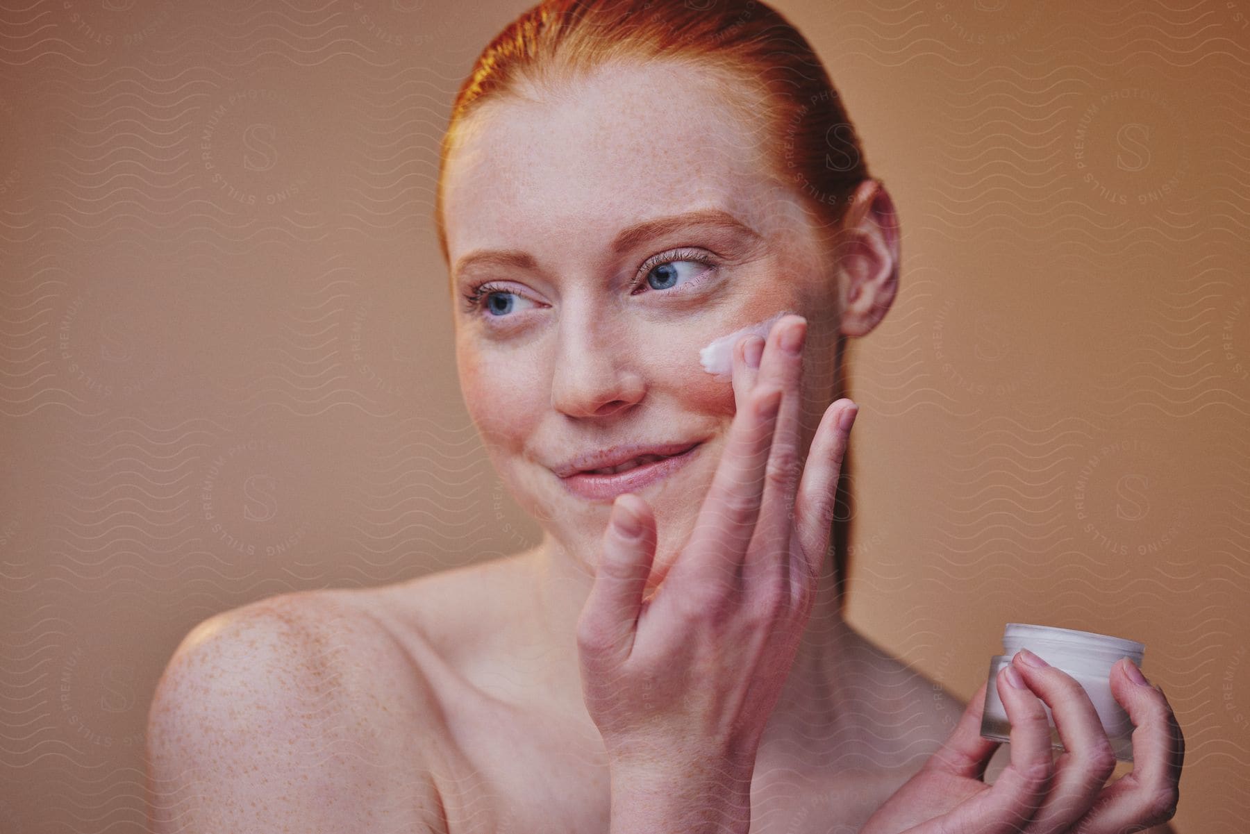 Young woman smiles and looks to her side as she applies skin cream to her face