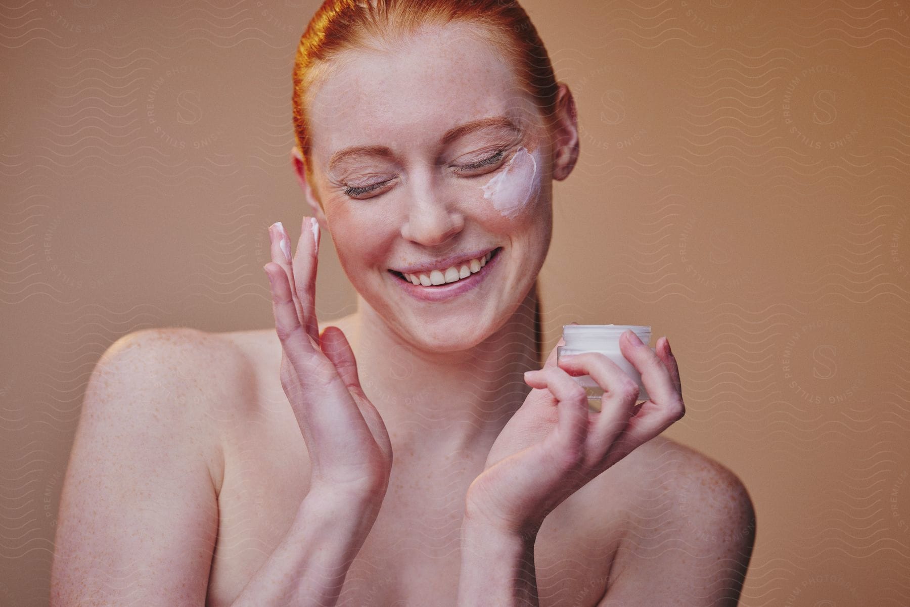 A woman smiles while rubbing face cream on her cheek.