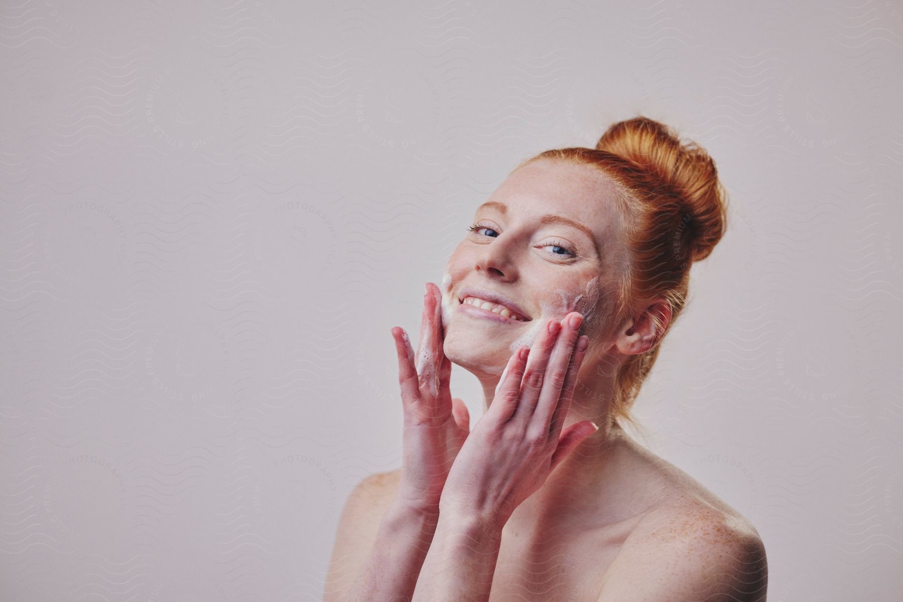 Smiling woman with red hair in a bun applying facial cleanser on her cheeks.