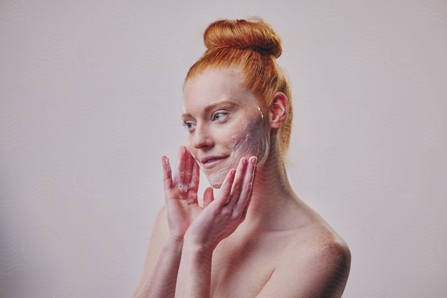 Woman with red hair up in a bun applying facial cleanser, looking to the side against a pale background.