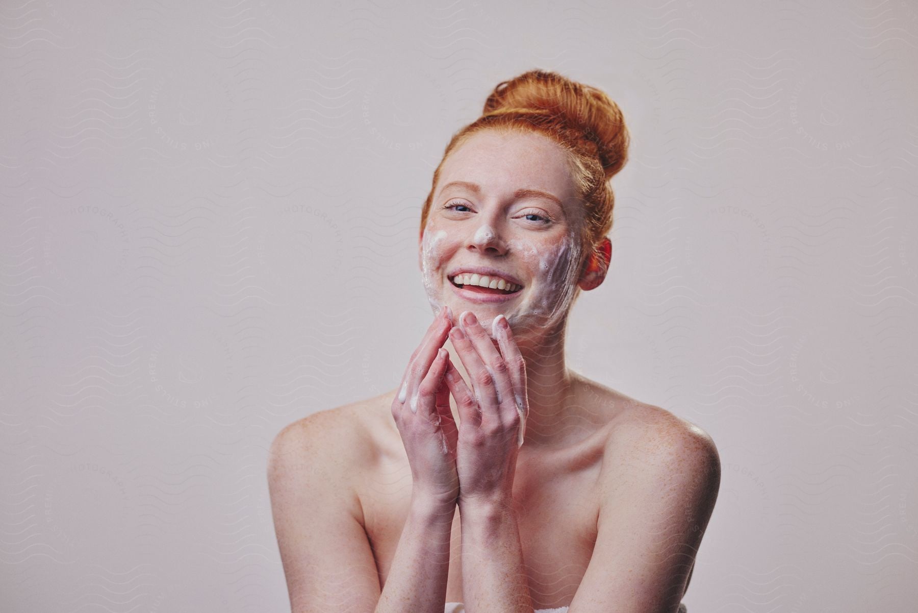 A woman with orange hair has a white facial cream on her face and she is smiling while her hands are on her chin