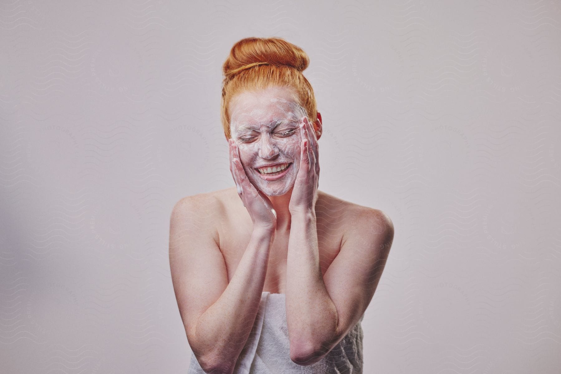 A woman with a bath towel and soap on her face smiles, her eyes closed and her hands flat against her face.