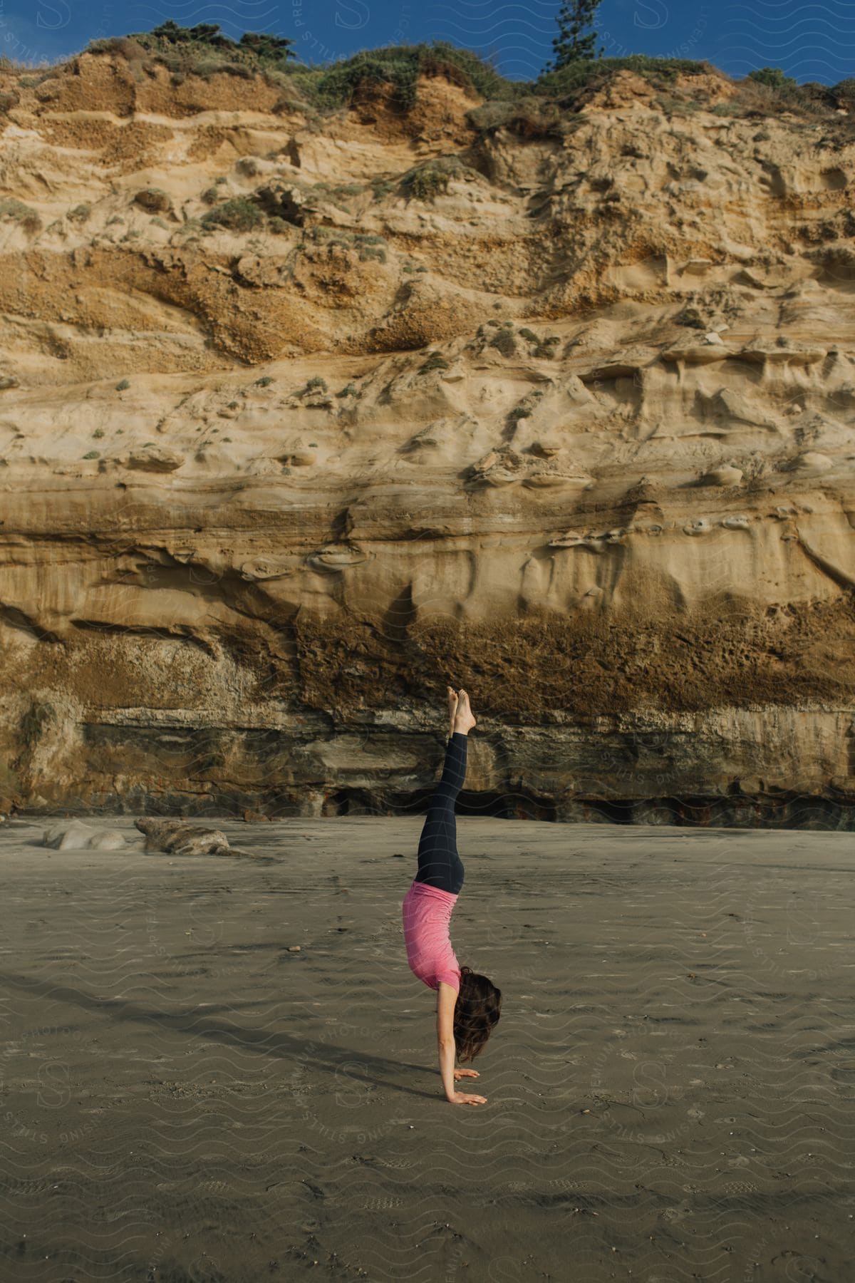 Woman performing a handstand on a sandy beach with a rugged cliff in the background.