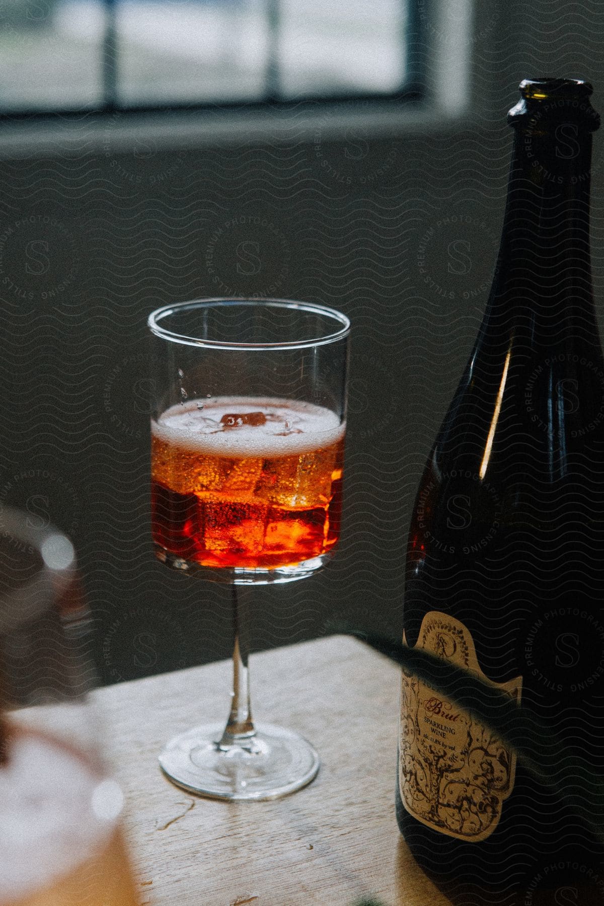 Glass of drink with ice and foam next to a dark bottle on a wooden table.