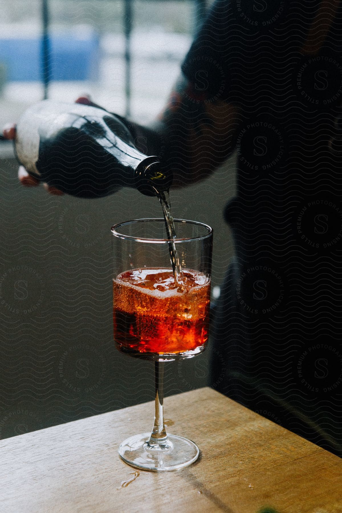 Man pouring a dark-colored beverage into a stemmed glass on a wooden surface.