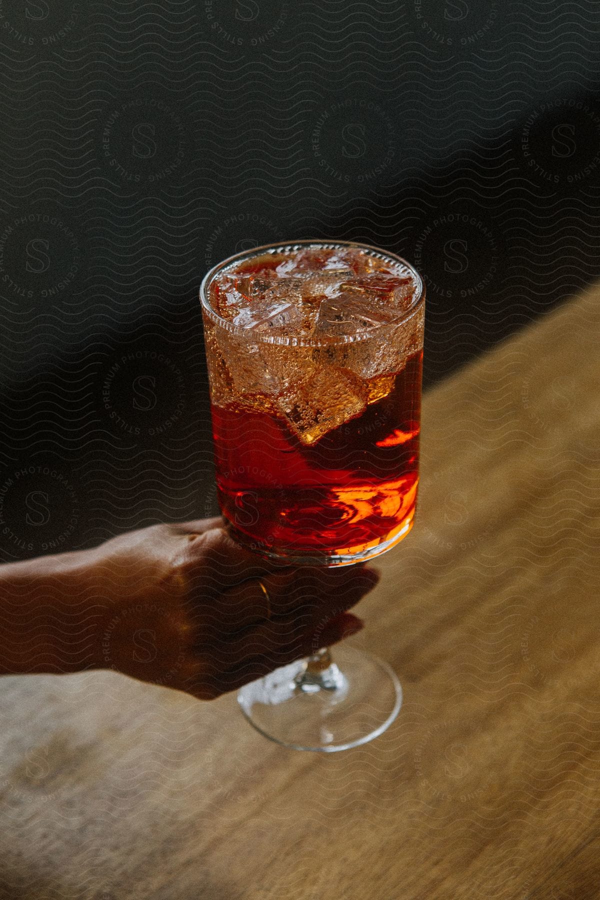 A person's hand holds a glass with ice cubes and a red liquid, on a brown wooden table.