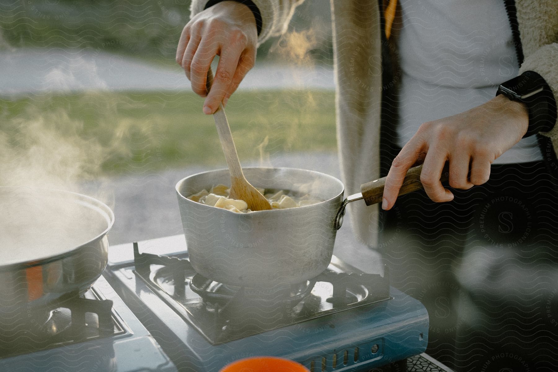 A person outdoors cooking on a blue portable stove and stirring one of two pans with a wooden spoon.