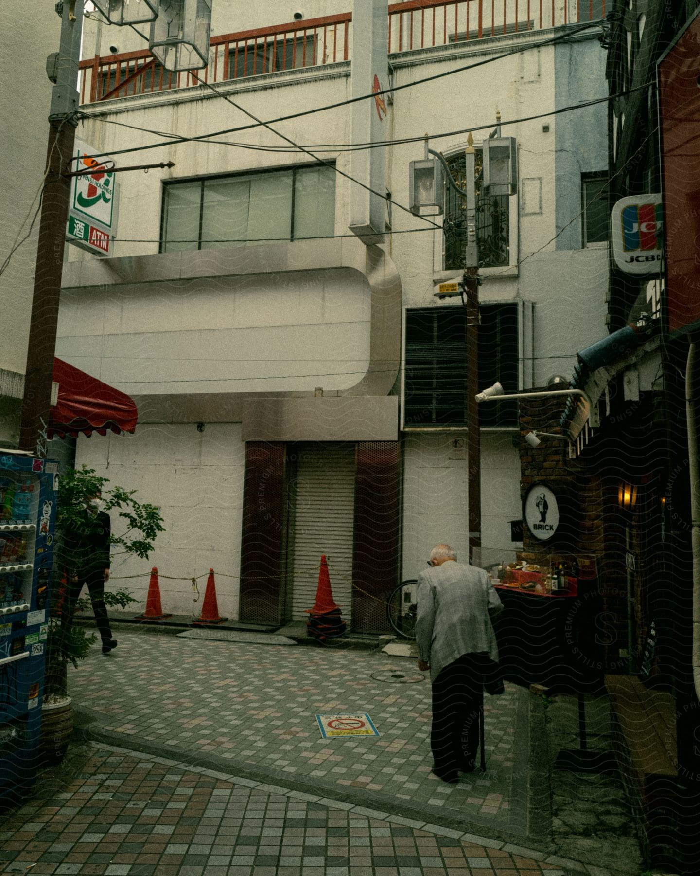 Elderly man walking in a quiet urban alley with shopfronts and a vending machine.