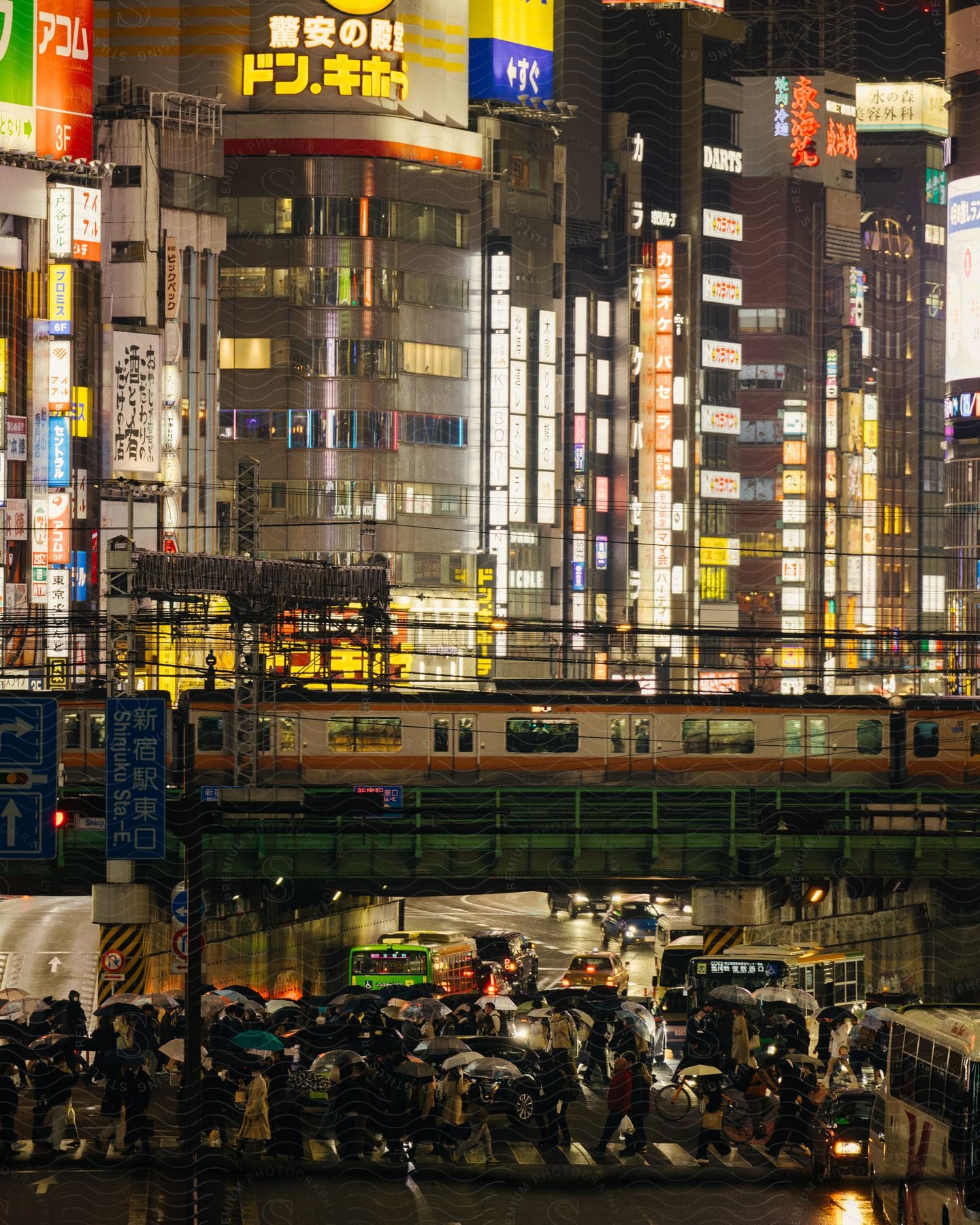 A busy urban scene at night, with skyscrapers illuminated by building lights and a train passing over a crowded street where people walk under umbrellas.