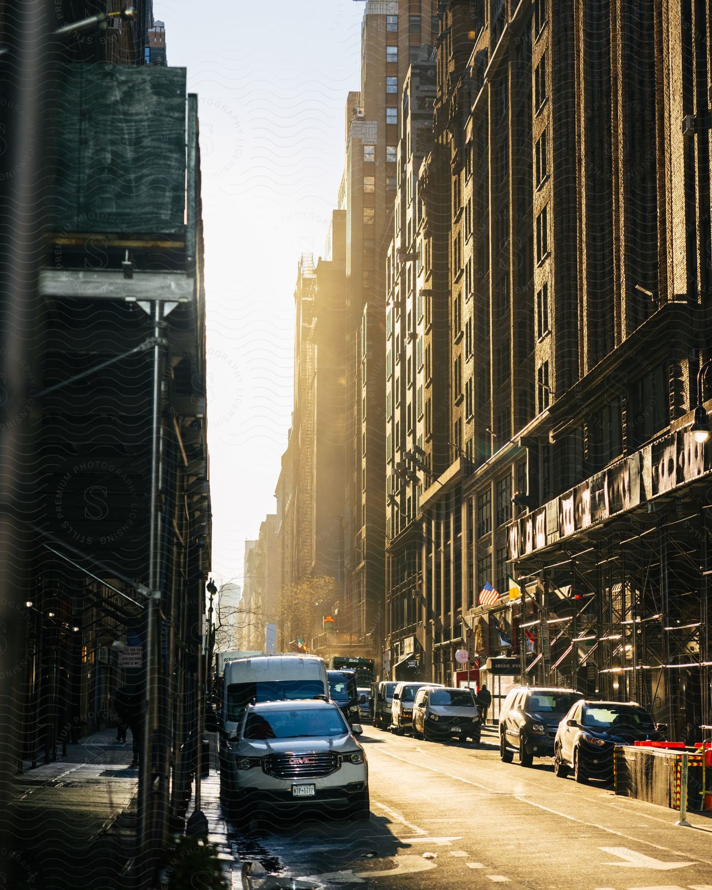 A narrow canyon of towering buildings casts long shadows as the sun dips low, bathing parked cars and pedestrians under awnings in a warm glow.