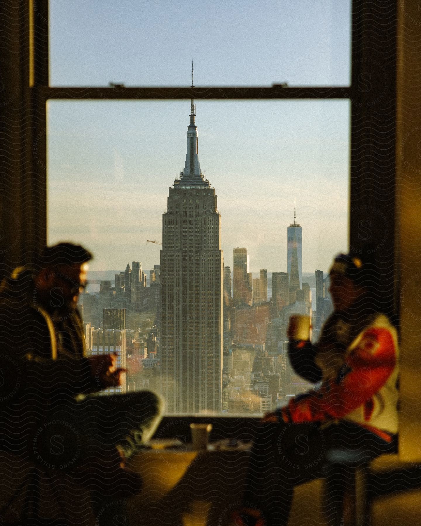 Two people sit next to a large window while drinking coffee and the Empire State building is visible.