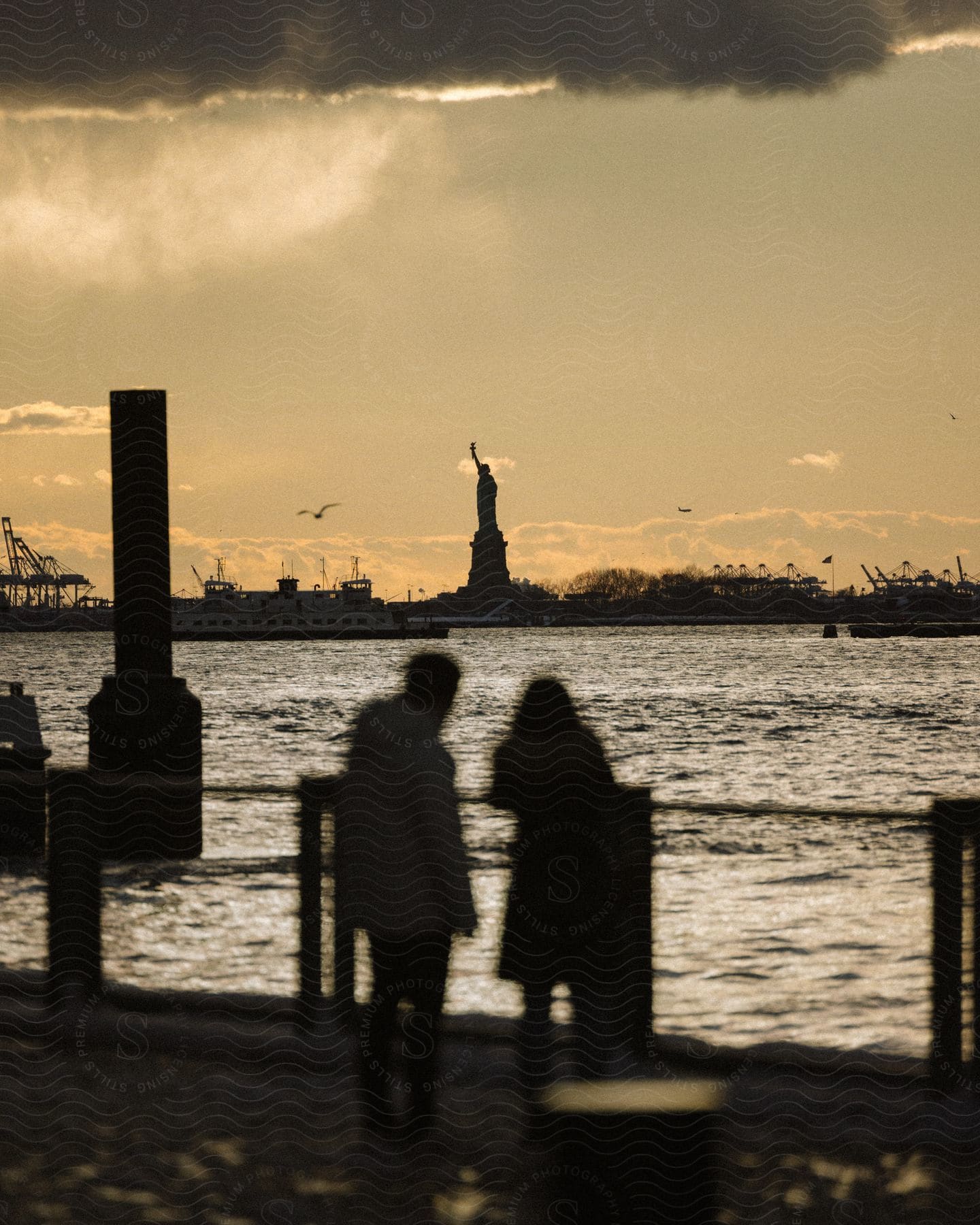 Silhouette of two people on the edge of a lake with a view of the Statue of Liberty in the background against a yellowish late afternoon sky