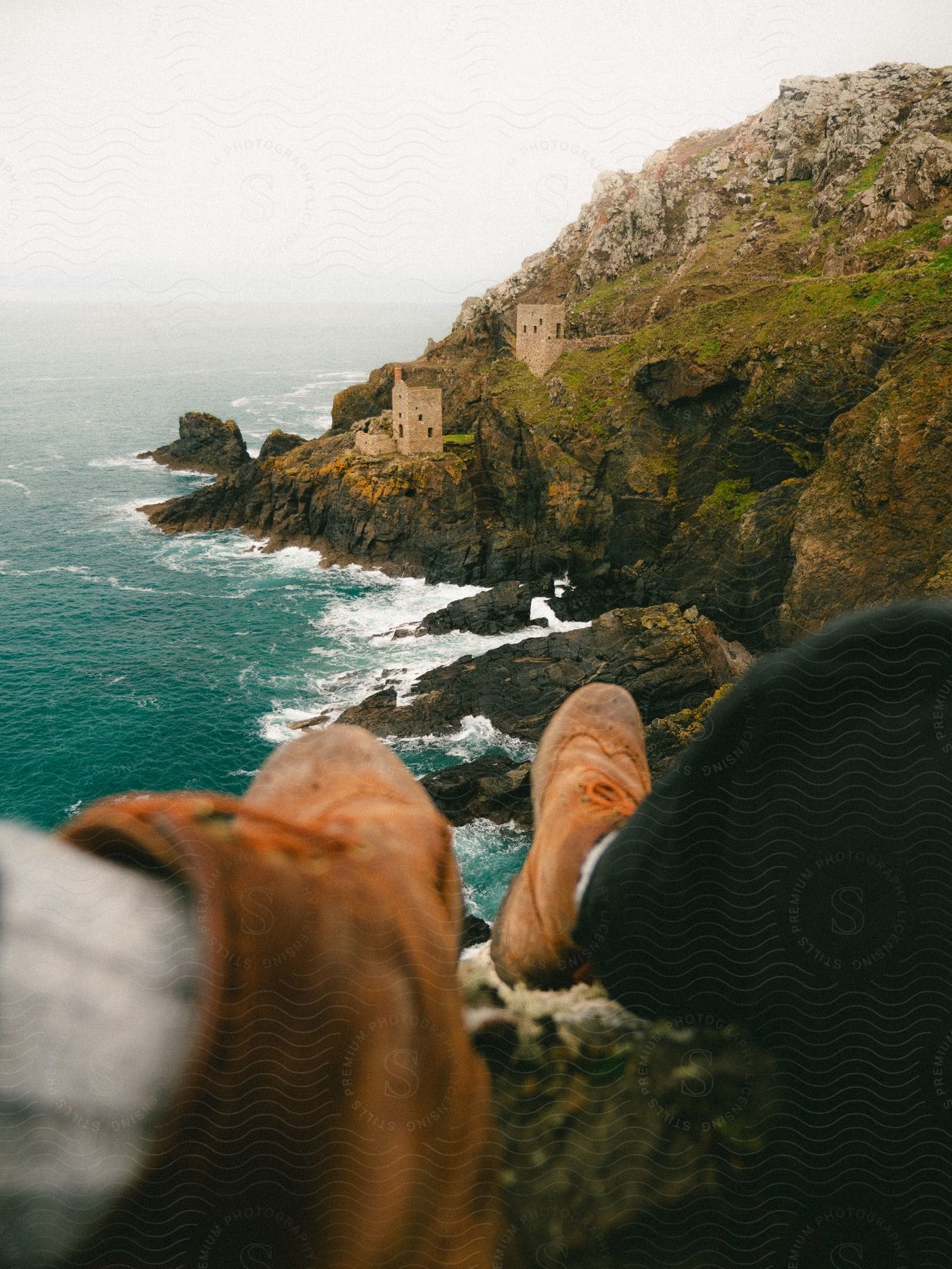 A person's boots dangle precariously over a rocky beach, with distant buildings perched on a mountainside.