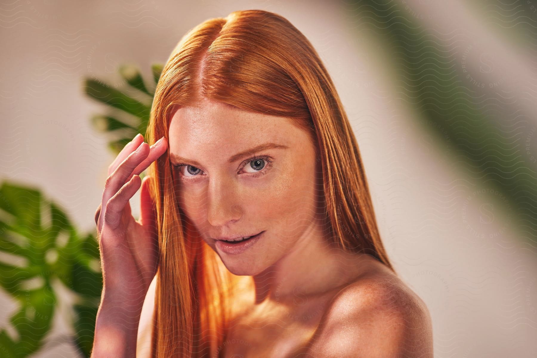 A young redheaded woman with light eyes looking straight ahead with a small smile and she has one hand on her forehead