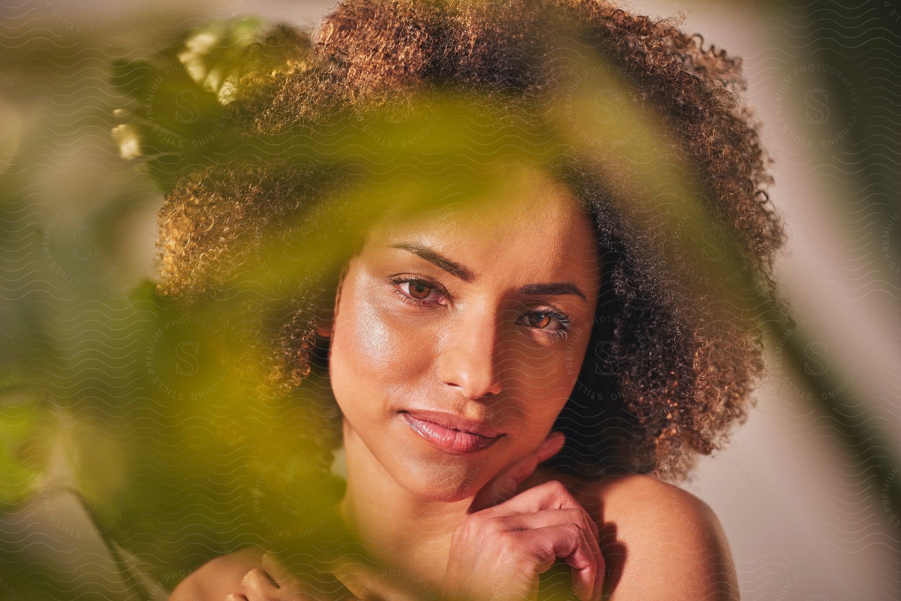 Portrait of a woman with curly hair with a hand on her face against a white background and blurry green leaves