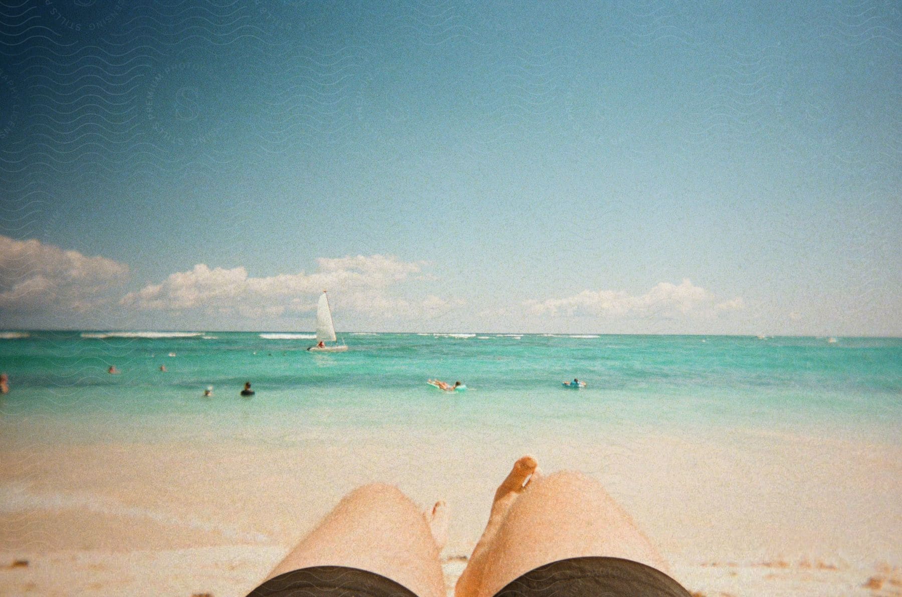 A water color painting of a beach with people and boats in the water, and a person with brown shorts from the legs down laying on the beach.