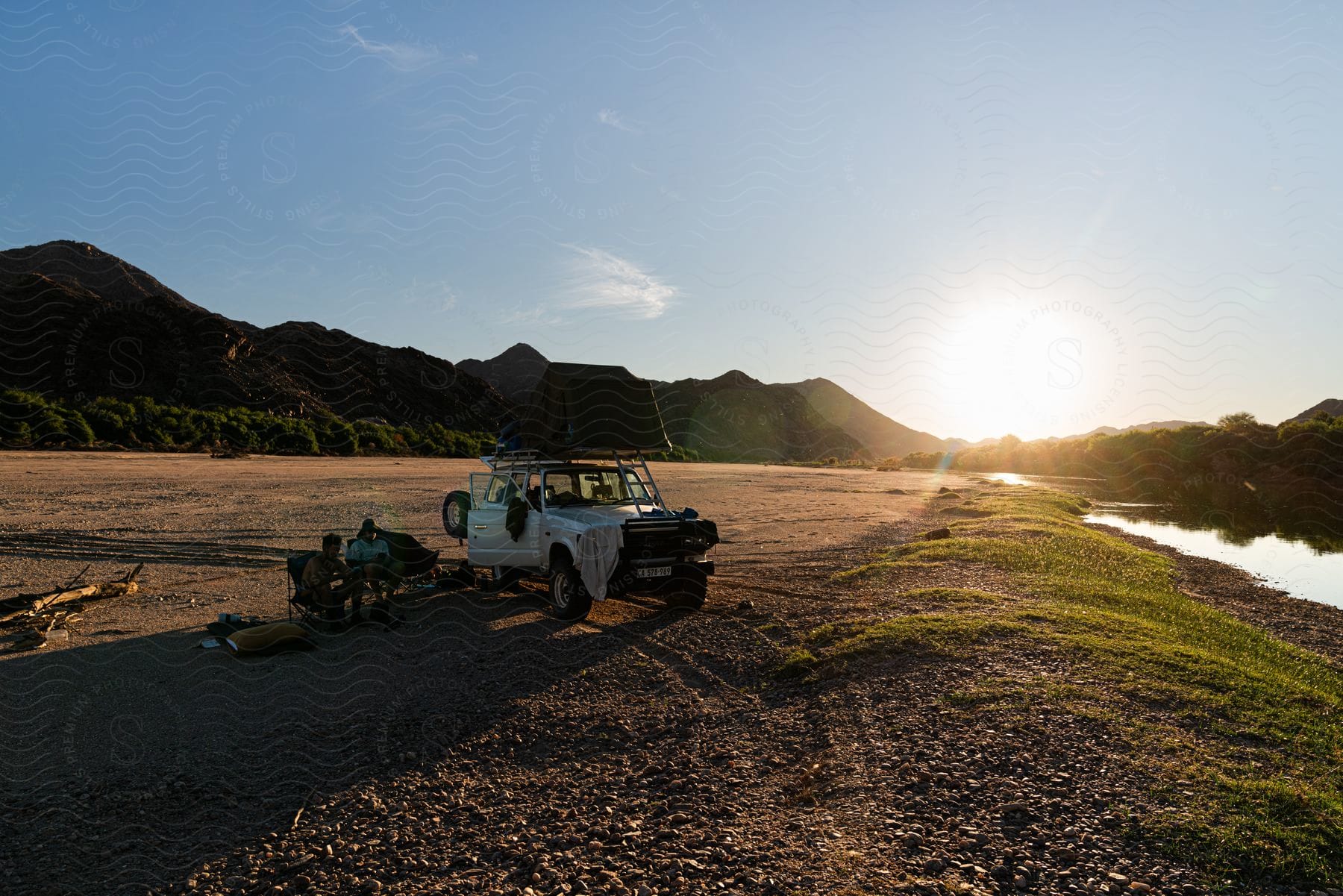 Jeep stopped on a barren ground with two people sitting on the side and the sunset on the horizon with the silhouette of mountains