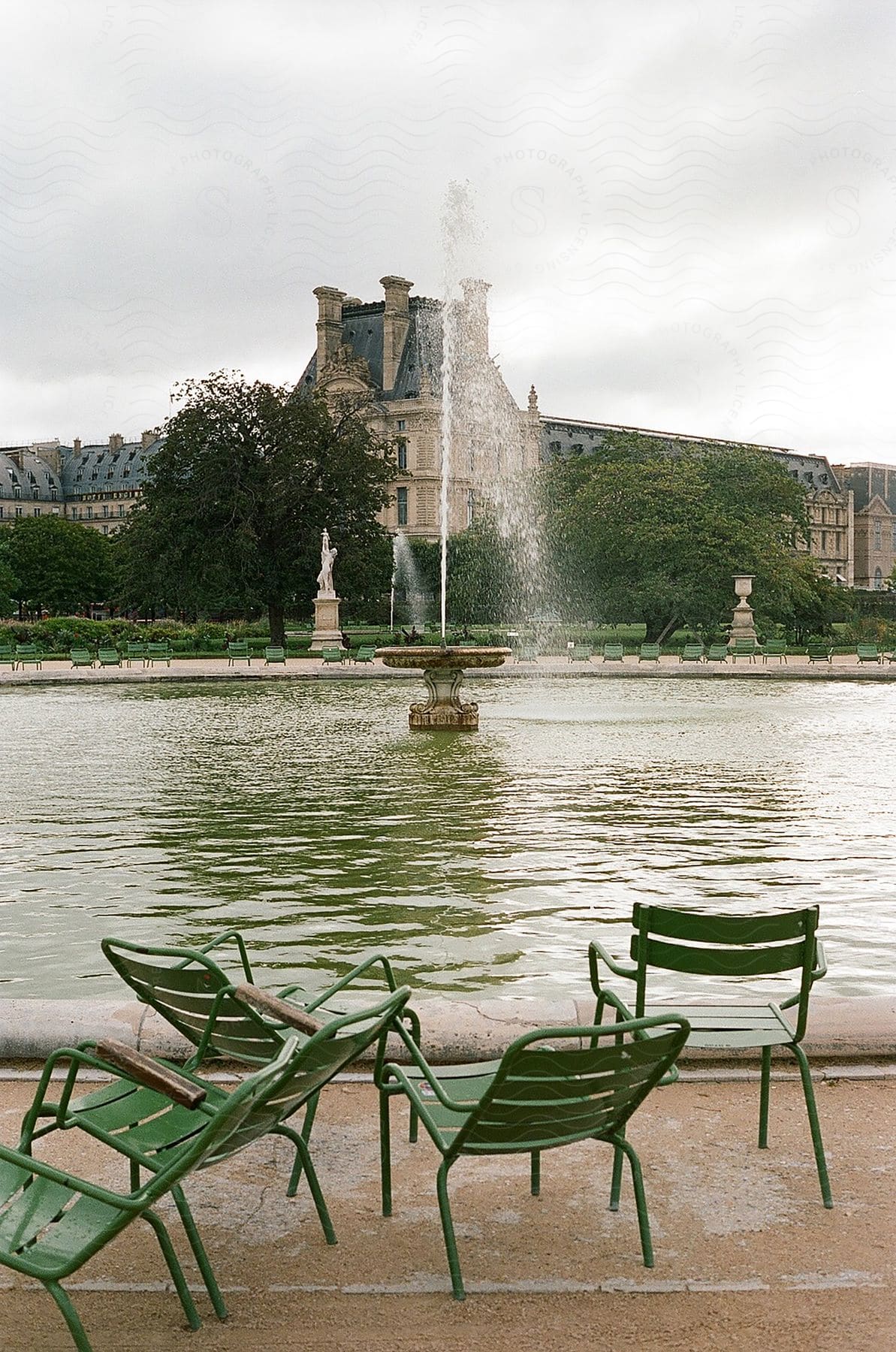 An old English-style building overlooks a pond with a central fountain spraying water, while green chairs rest on the cement foreground.