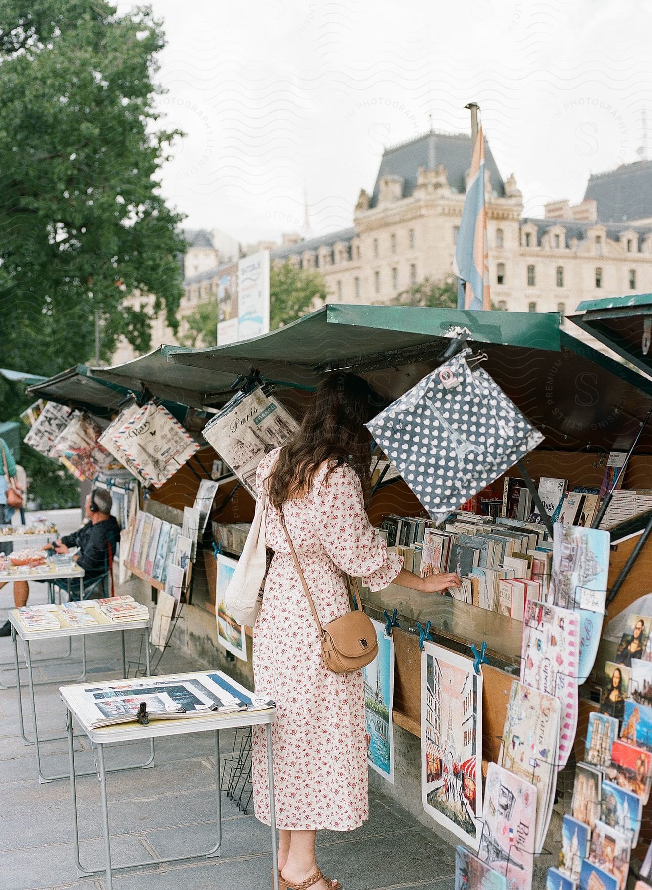 Woman in floral dress looking at books at an outdoor stall full of magazines and postcards in a city square.