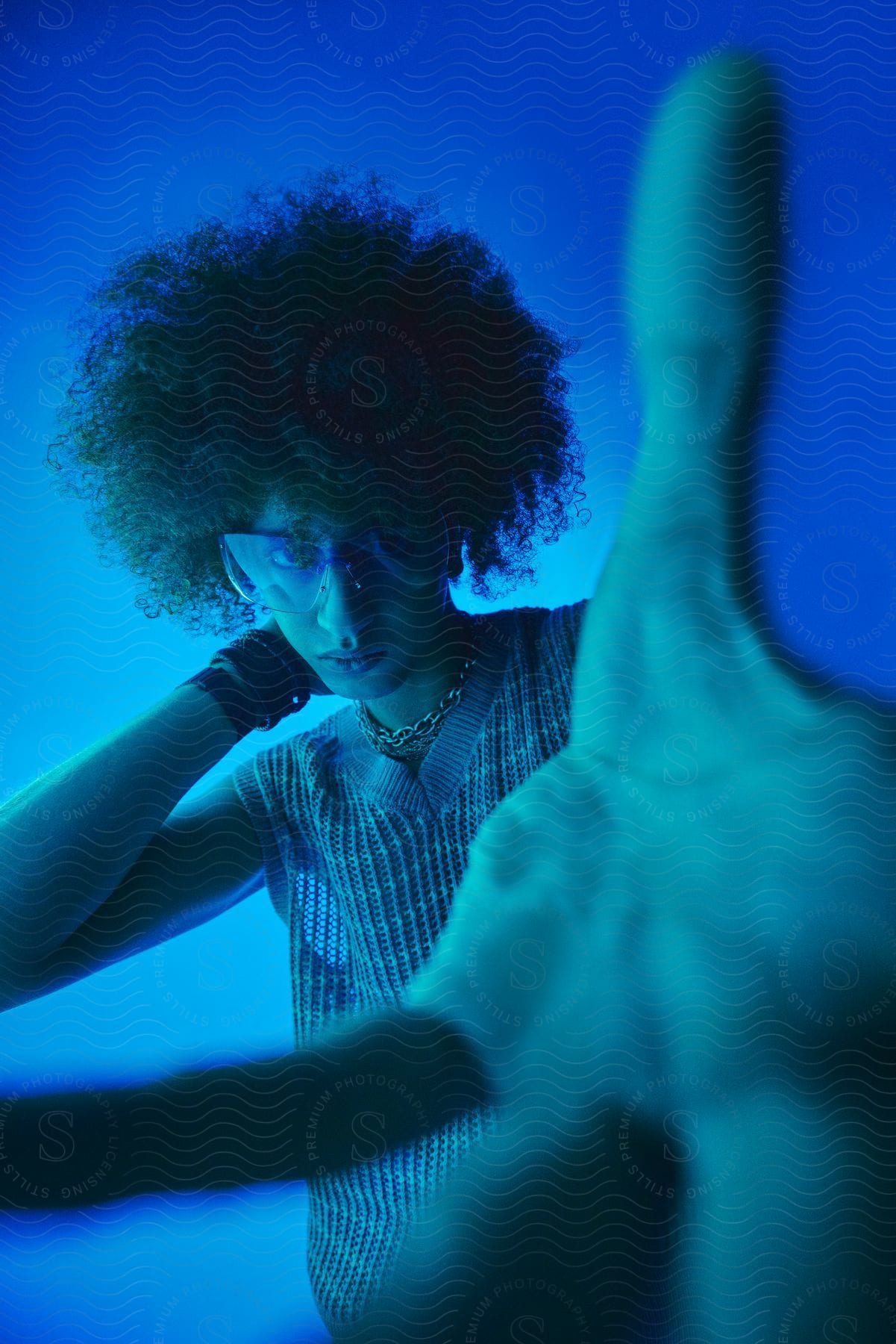 A woman with frizzy hair, wearing glasses, bracelets, a necklace, and a blue vest, poses by reaching out her hand in the blue-lit studio