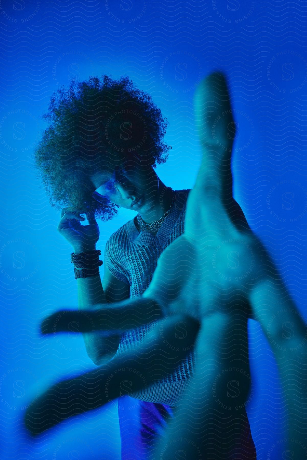 A person posing in a room, illuminated by a blue light, with an outstretched hand blurred in the foreground.