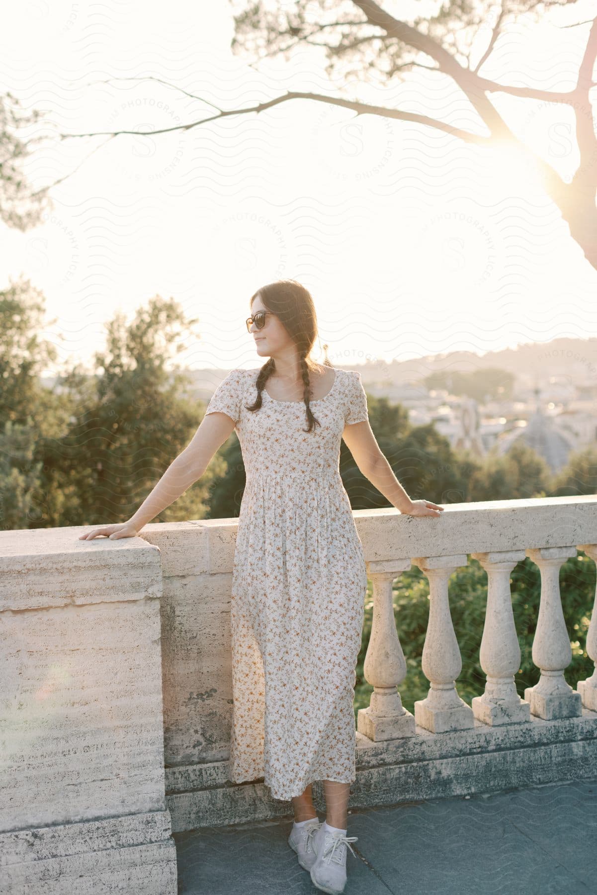 A woman in a floral dress, sunglasses perched on her nose, takes in the city view with arms outstretched on a balcony railing.