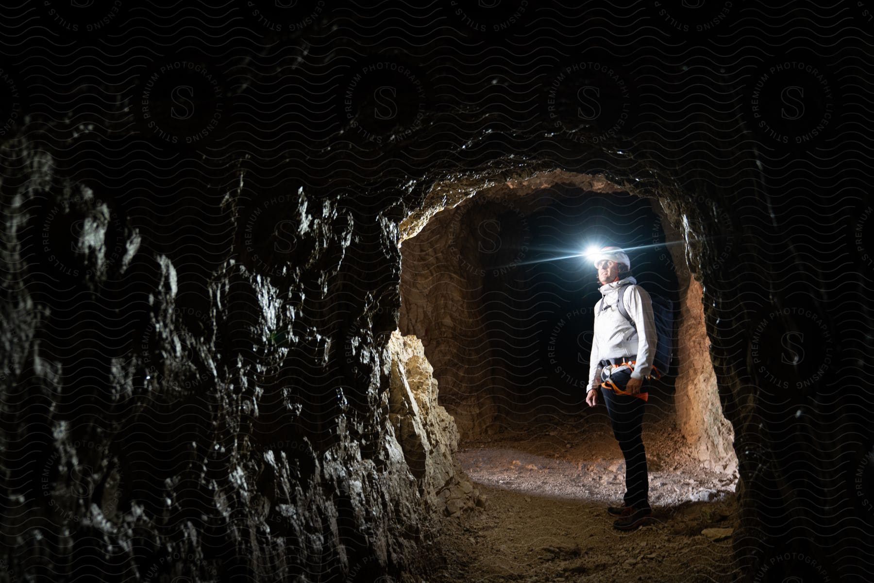 Man exploring a dark cave with a headlamp, wearing a white shirt, dark pants, and a backpack, standing in a tunnel illuminated by the headlamp light.