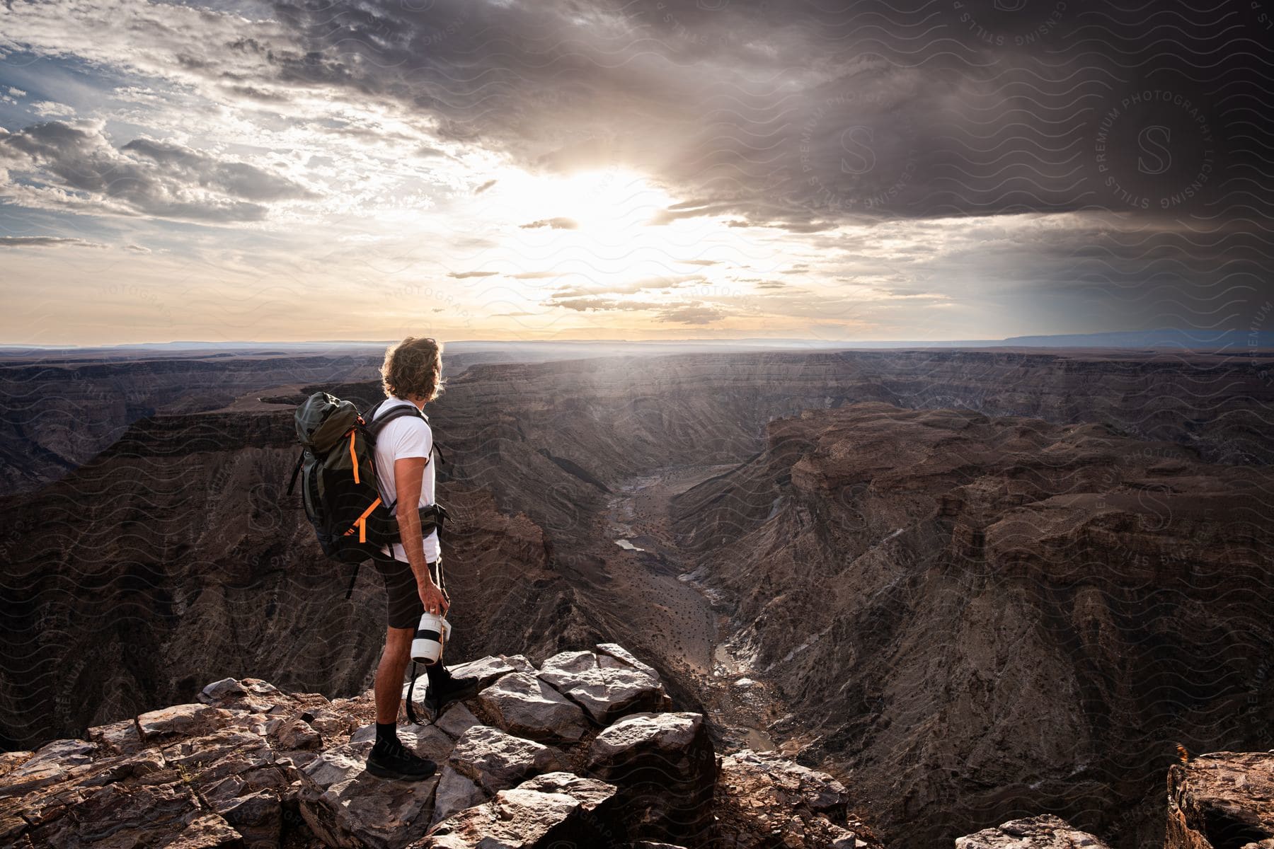 A hiker, wearing a white t-shirt, shorts, and a black backpack, watches the sun shining through dramatic clouds over a vast canyon