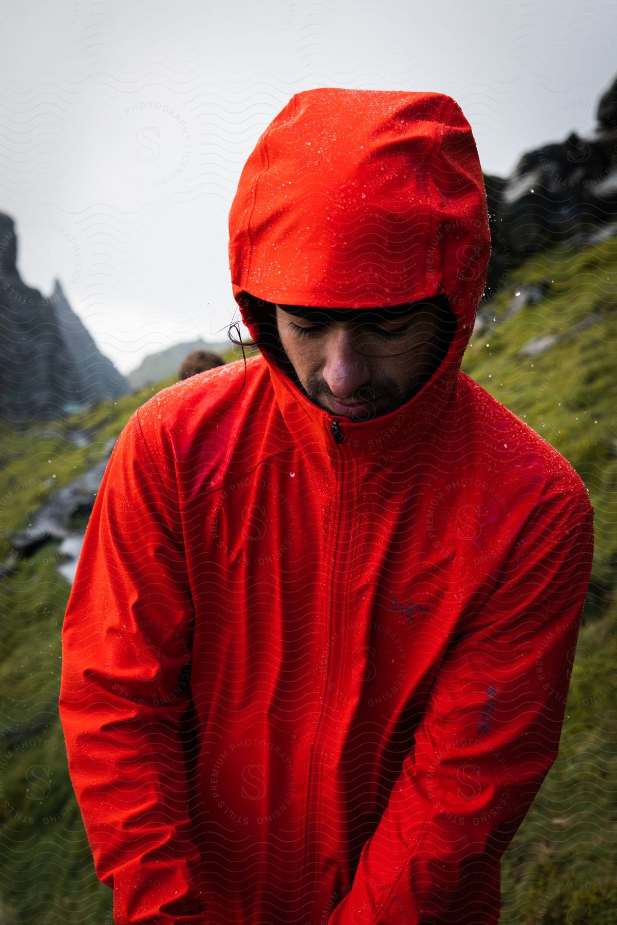 A man wearing a red hooded jacket looking down with jagged rocky ridge and green grass behind him