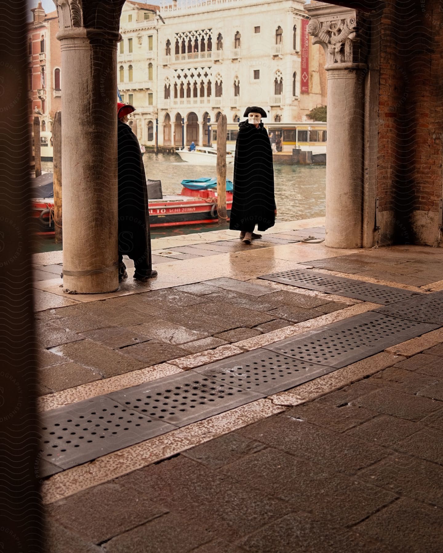 Masked Figures In Black Robes Stand At A Venetian Building Entrance, Gondolas Lining The Canal Behind Them