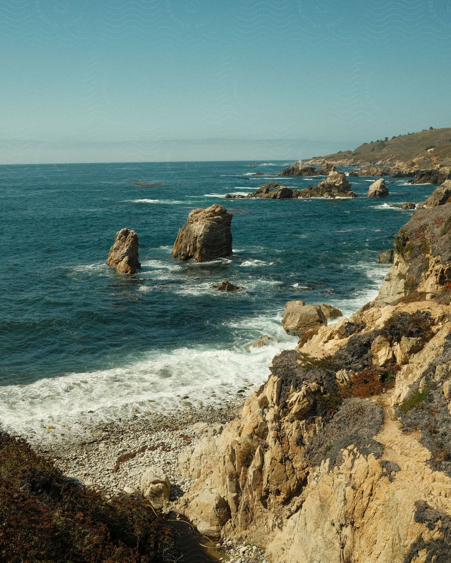 A coastal landscape with imposing rocks emerging from the rough ocean next to a rocky cliff.