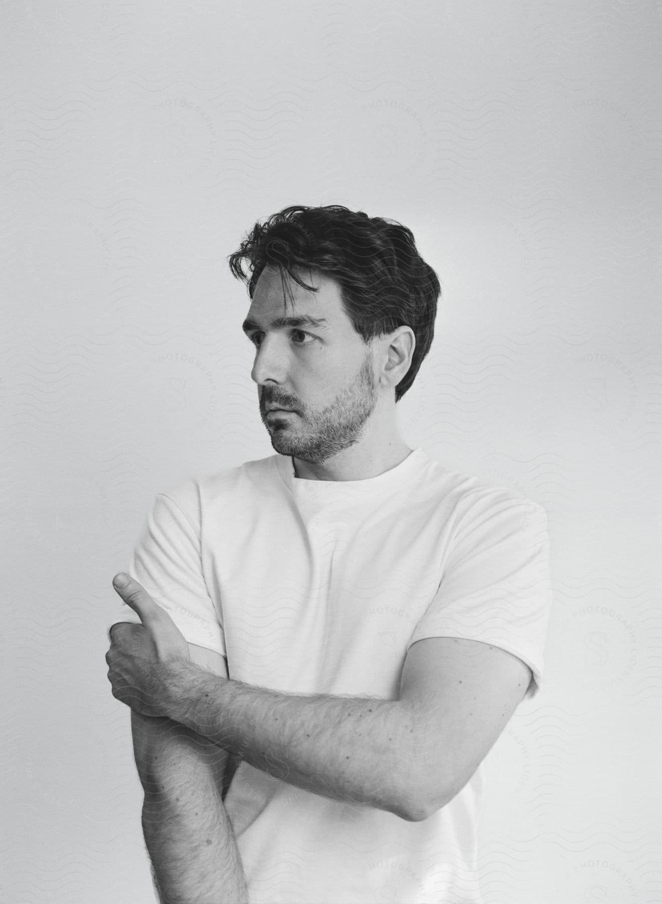 Bearded man looking to the side with one hand on his arm and wearing a white t-shirt indoors