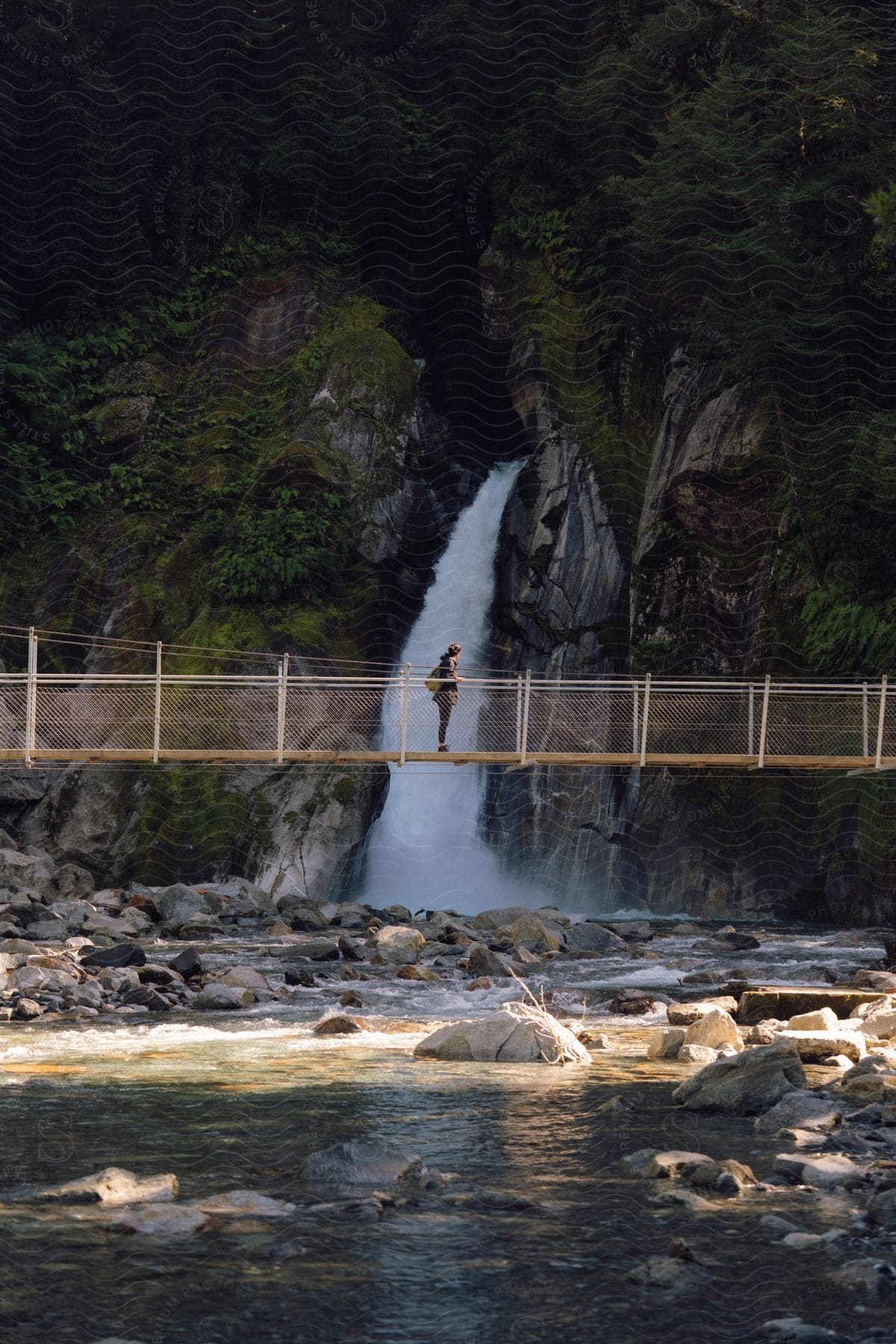 Woman standing on a suspension bridge over a creek admiring a waterfall flowing from the canyon