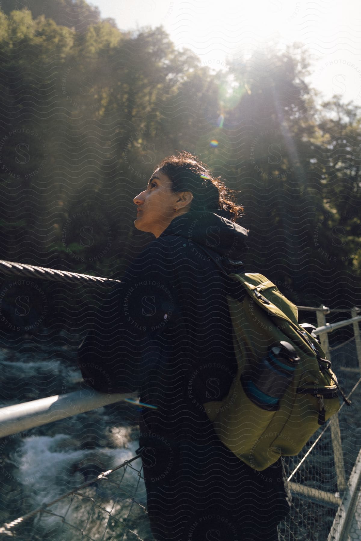Woman with a backpack standing on a suspension bridge over a river, sunlight filtering through trees.