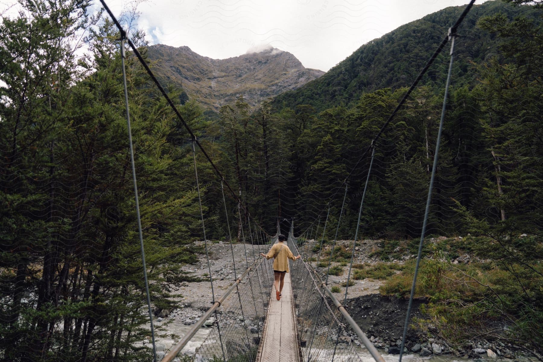 A person in a yellow shirt is walking on a wooden suspension bridge in a region with many trees