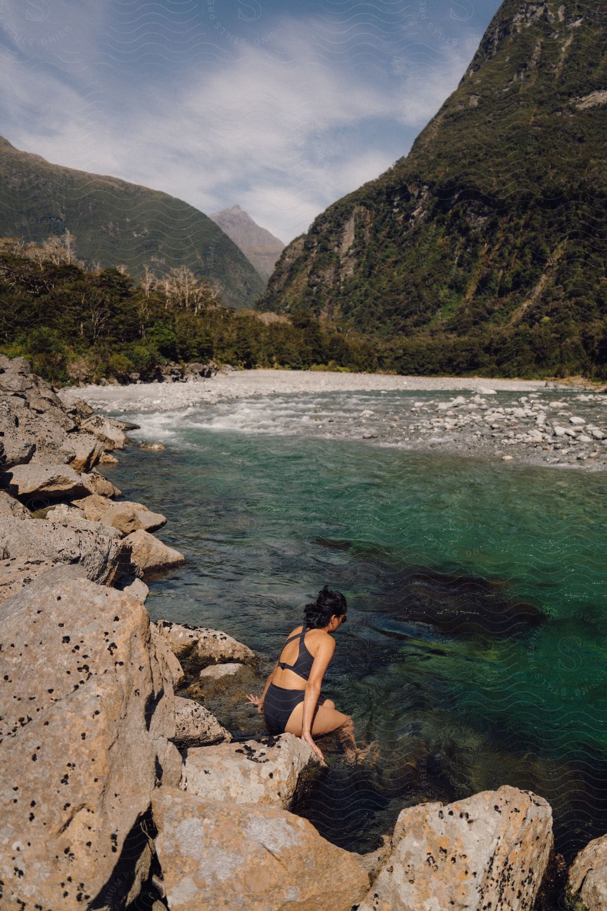 A person is entering a clear, turquoise river, surrounded by rocky terrain and green mountains.