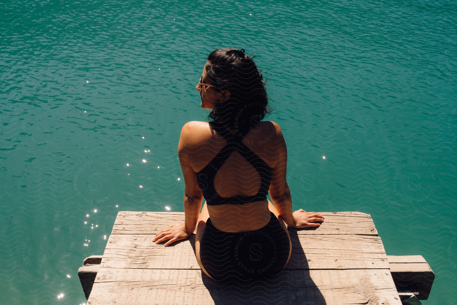 Woman in a bikini sitting on a wooden dock overlooking turquoise water.