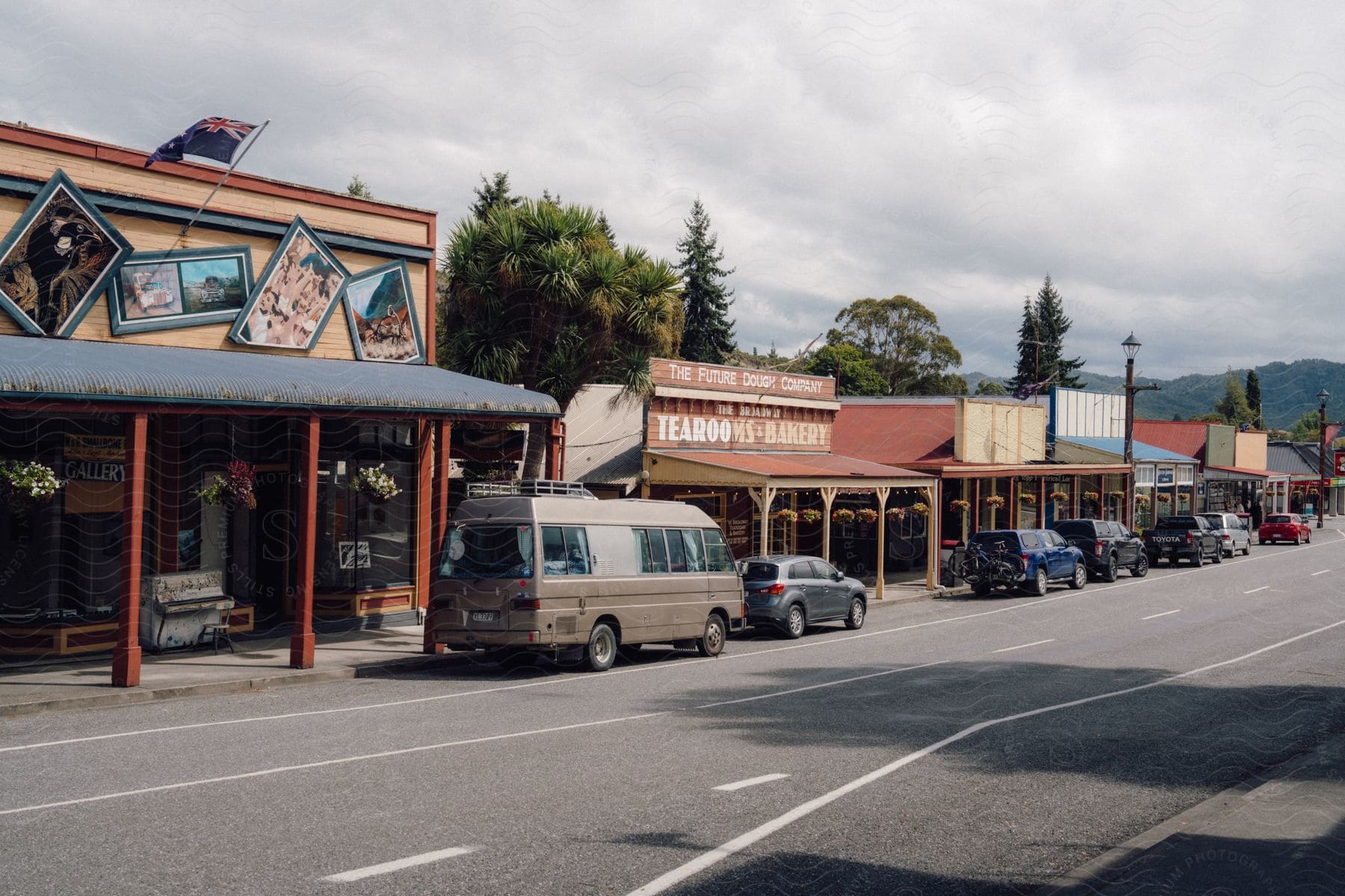 Cars are parked on the street next to a bakery and other small-town businesses on a cloudy day.