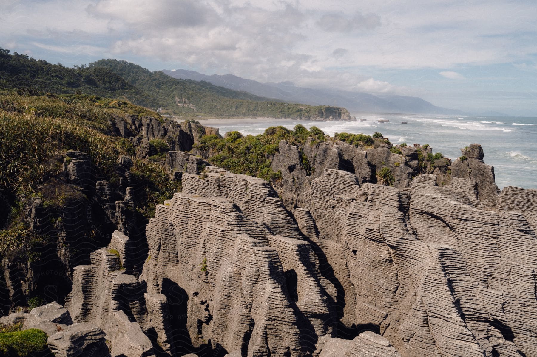 Rocky coastal landscape featuring weathered stone formations overlooking a lush coastline with distant mountains.