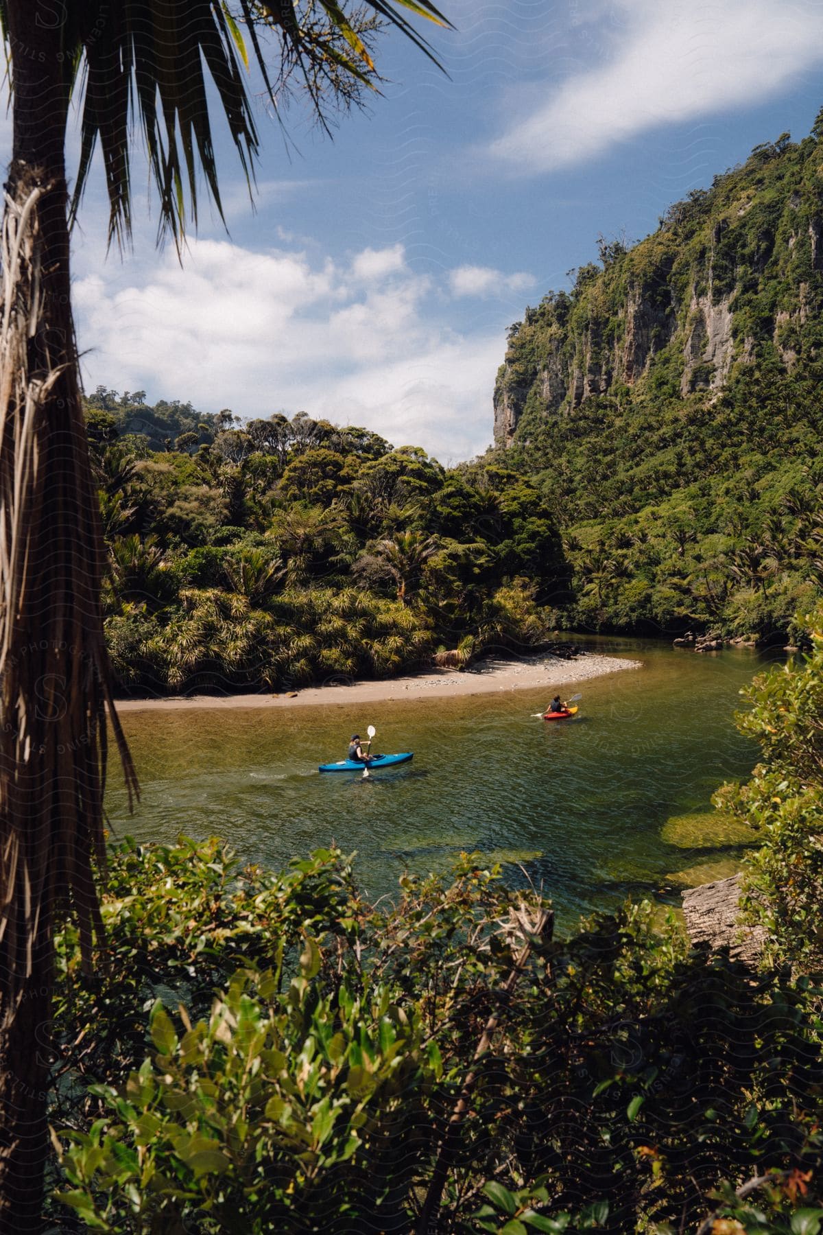 Two individuals are canoeing through the water near a tropical beach, with a high rocky cliff visible in the distance.