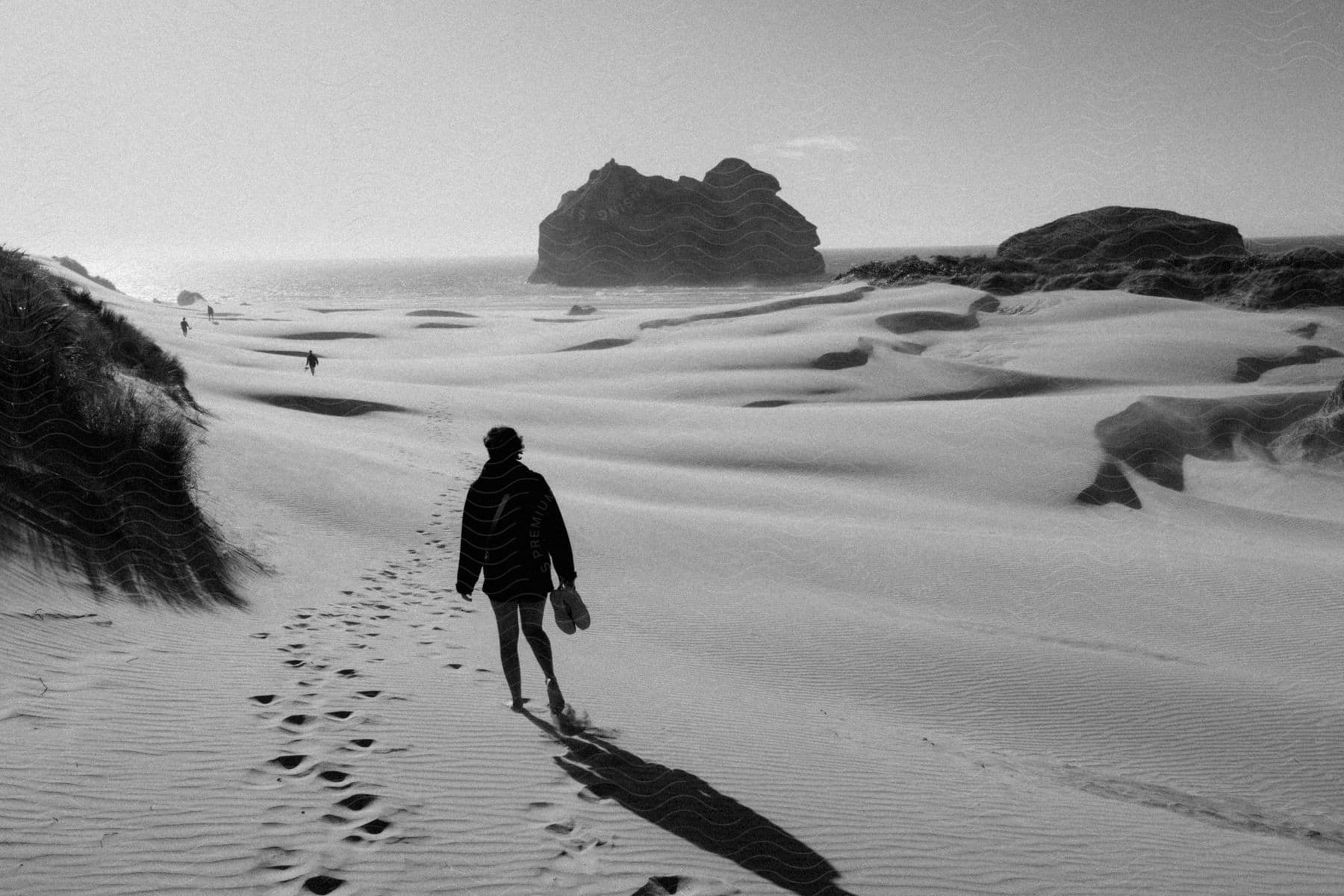 Black and white of a woman walking barefoot in the sand near the ocean on a sunny day.