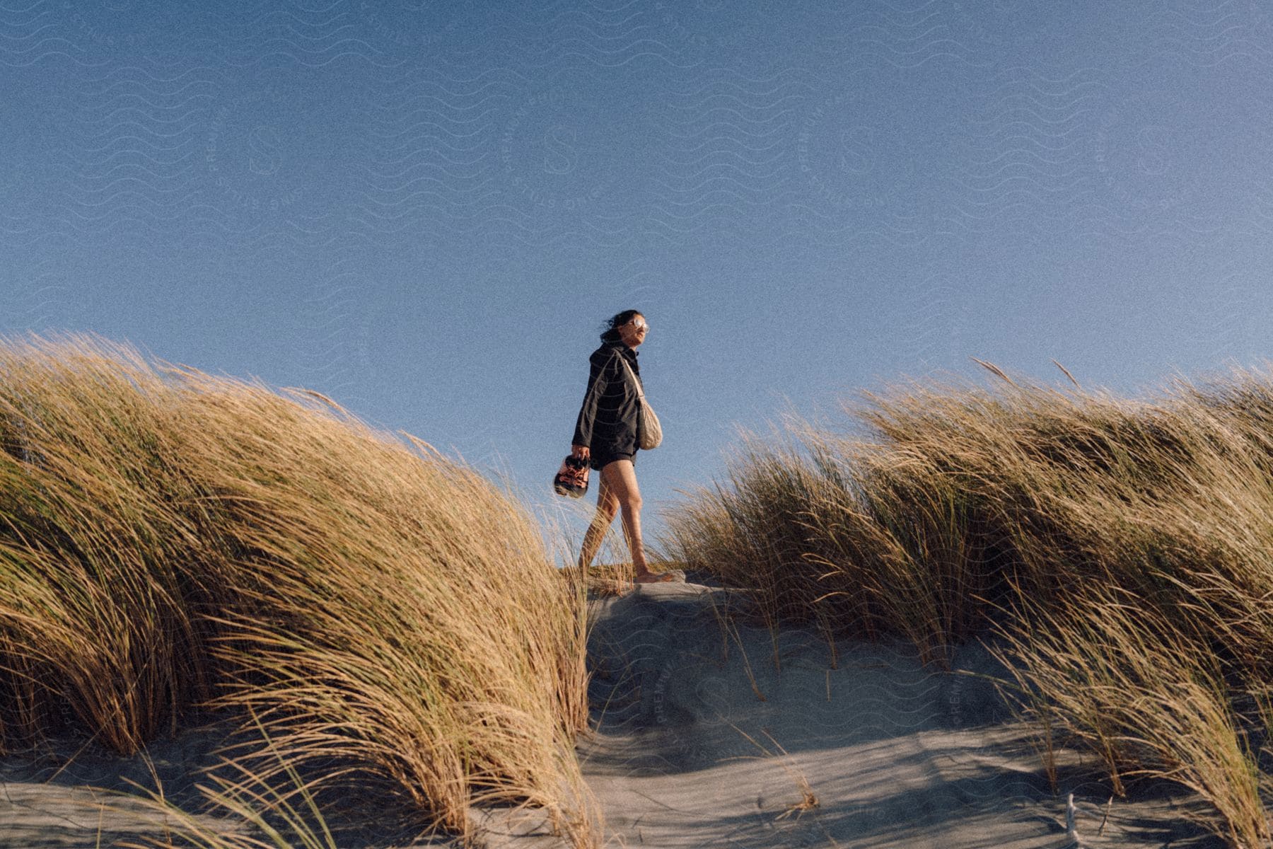 A woman walks barefoot in the sand next to tall beach grass on a sunny day.